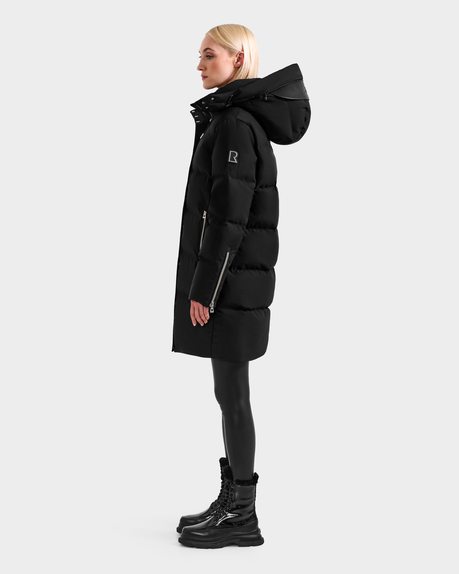 A woman with straight blonde hair models the Rudsak JOELLE H - 8125602 Black puffer coat with a hood, paired with black leggings and lace-up boots, standing in profile against a plain light gray background.
