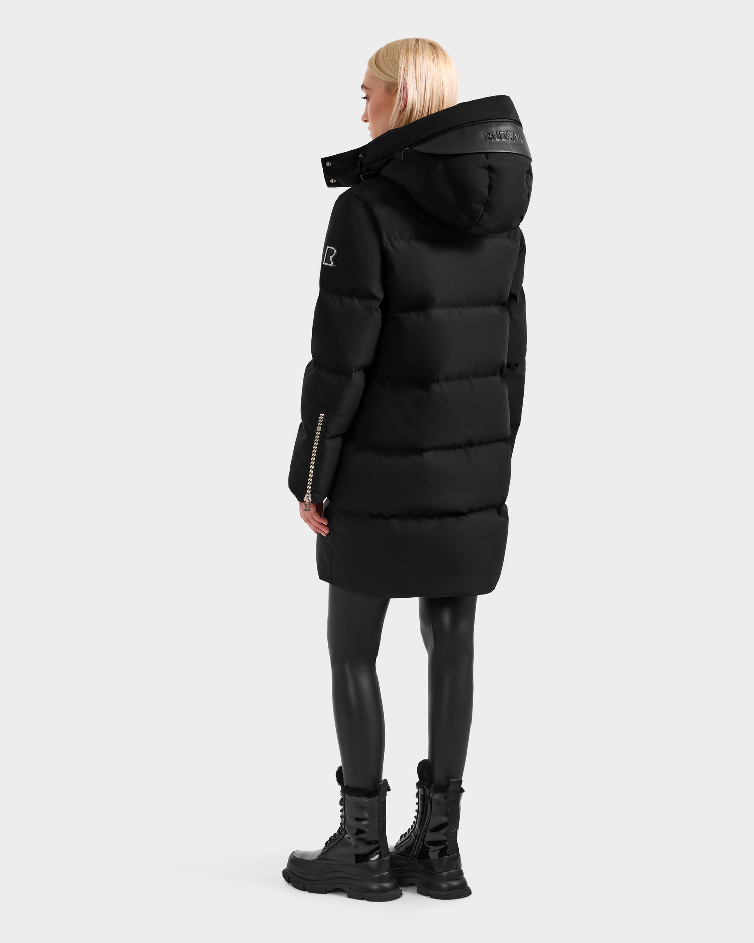 A person with light blond hair wears the Rudsak JOELLE H - 8125602 Black women’s down puffer, black leggings, and lace-up boots, standing slightly turned away against a plain light background.