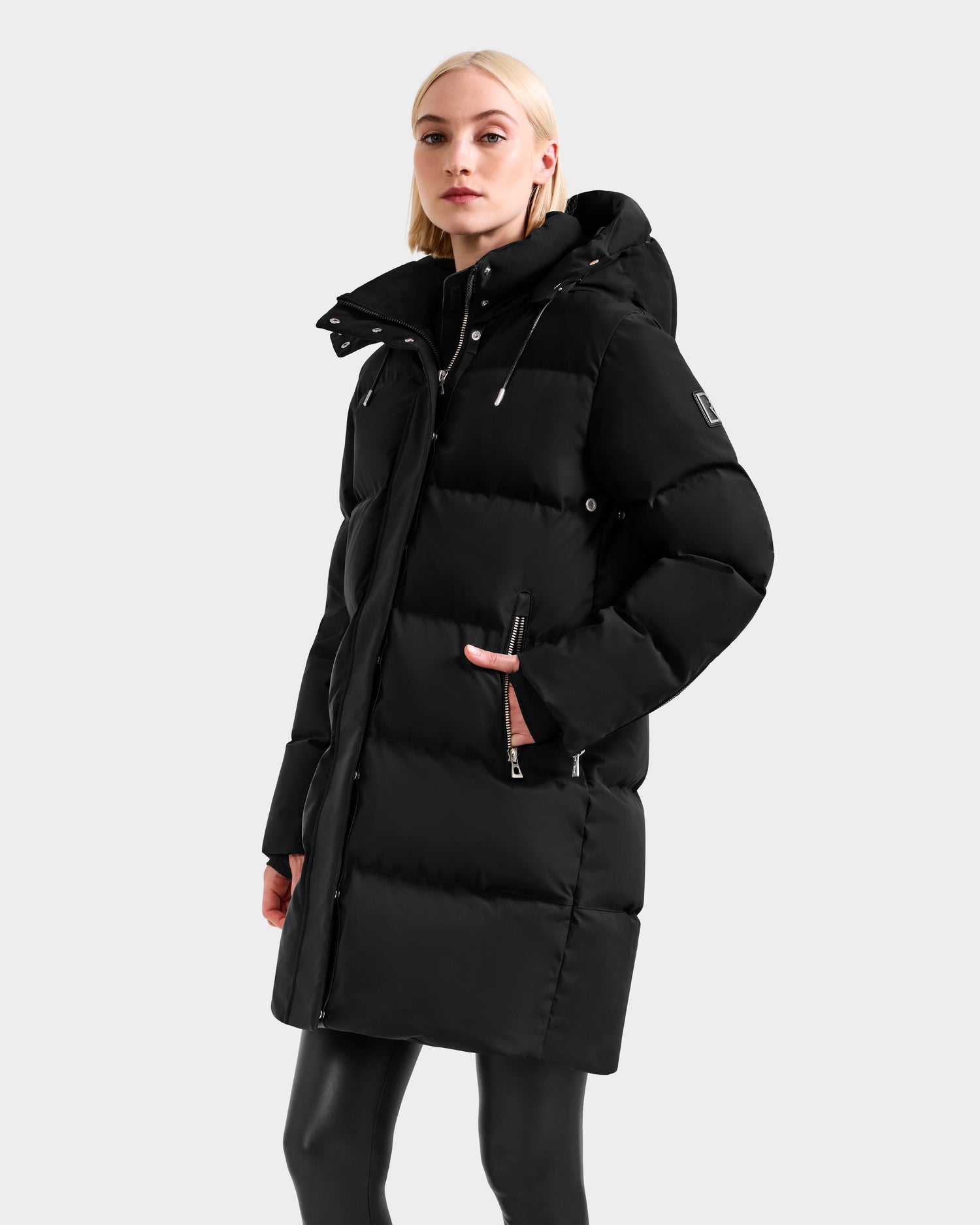 A woman with light hair models the Rudsak JOELLE H - 8125602 Black women’s down puffer coat, featuring a hood and zippered pockets, against a plain light gray background.