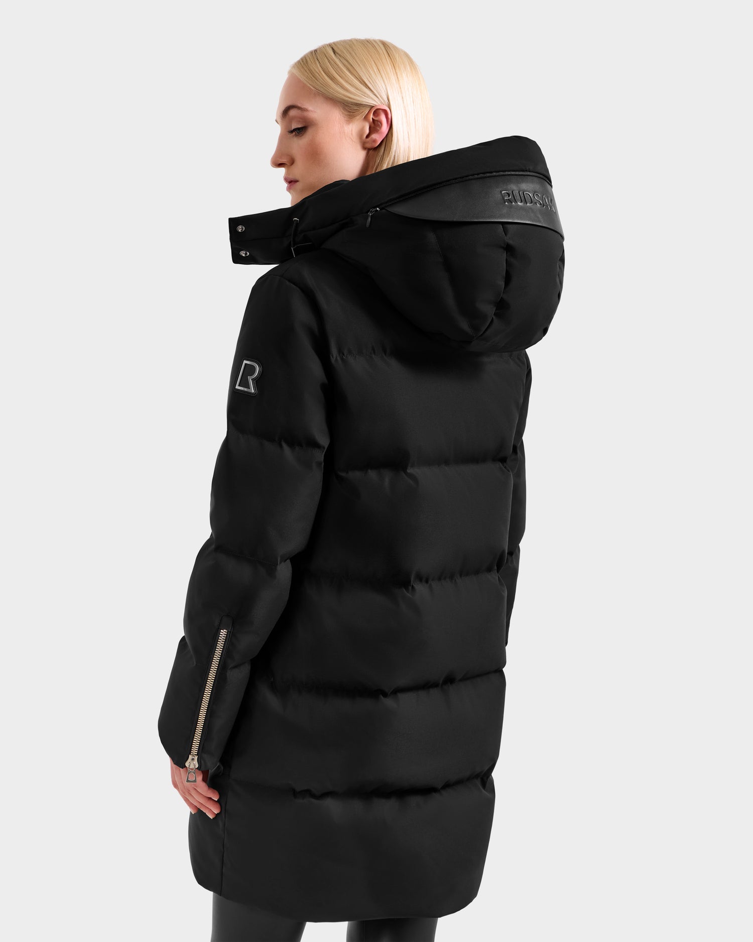 A person with straight blond hair wears the Rudsak JOELLE H - 8125602 Black women’s down puffer, featuring a long hooded design, a sleeve zipper, and visible logo details on the hood and upper arm, standing turned away at an angle.
