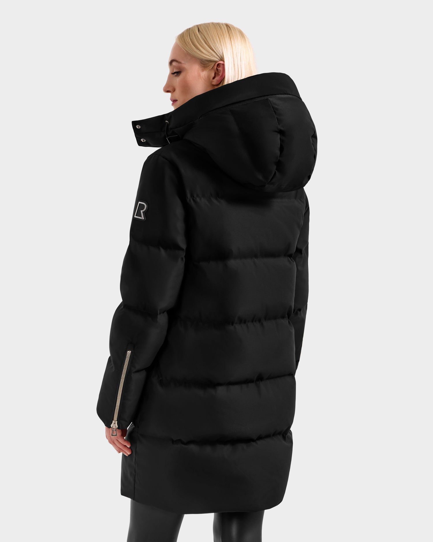 A woman with straight blonde hair, turned away from the camera, wears the Rudsak JOELLE H - 8125602 Black women’s down puffer coat with a hood. The water-resistant coat features a sleeve logo and zipper wrist detail, set against a light gray background.