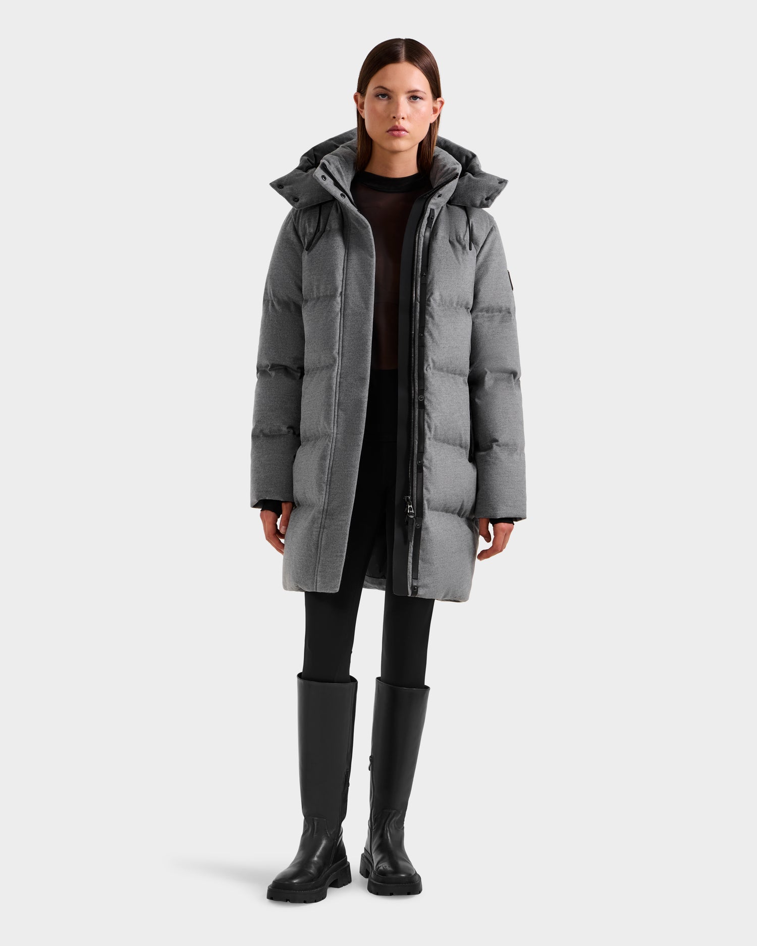 A woman wearing the Rudsak JOELLE W - 8125567 Heather Charcoal down puffer coat with a hood, black leggings, and knee-high boots stands facing forward against a white background—a must-have for cold weather.