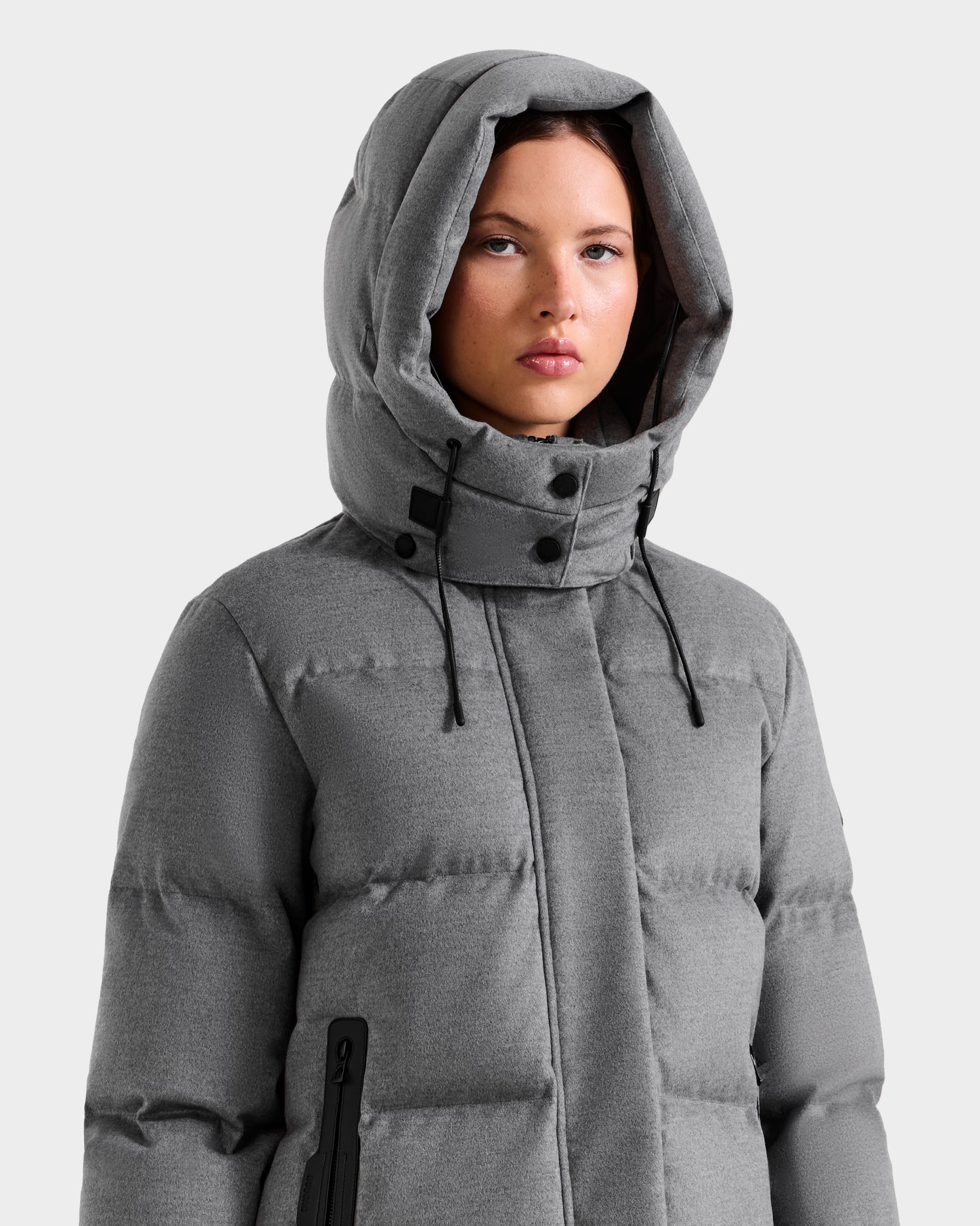 A woman wears the Rudsak JOELLE W - 8125567 Heather Charcoal down puffer jacket with a large hood and black zipper accents, standing against a light background—a must-have for cold weather.