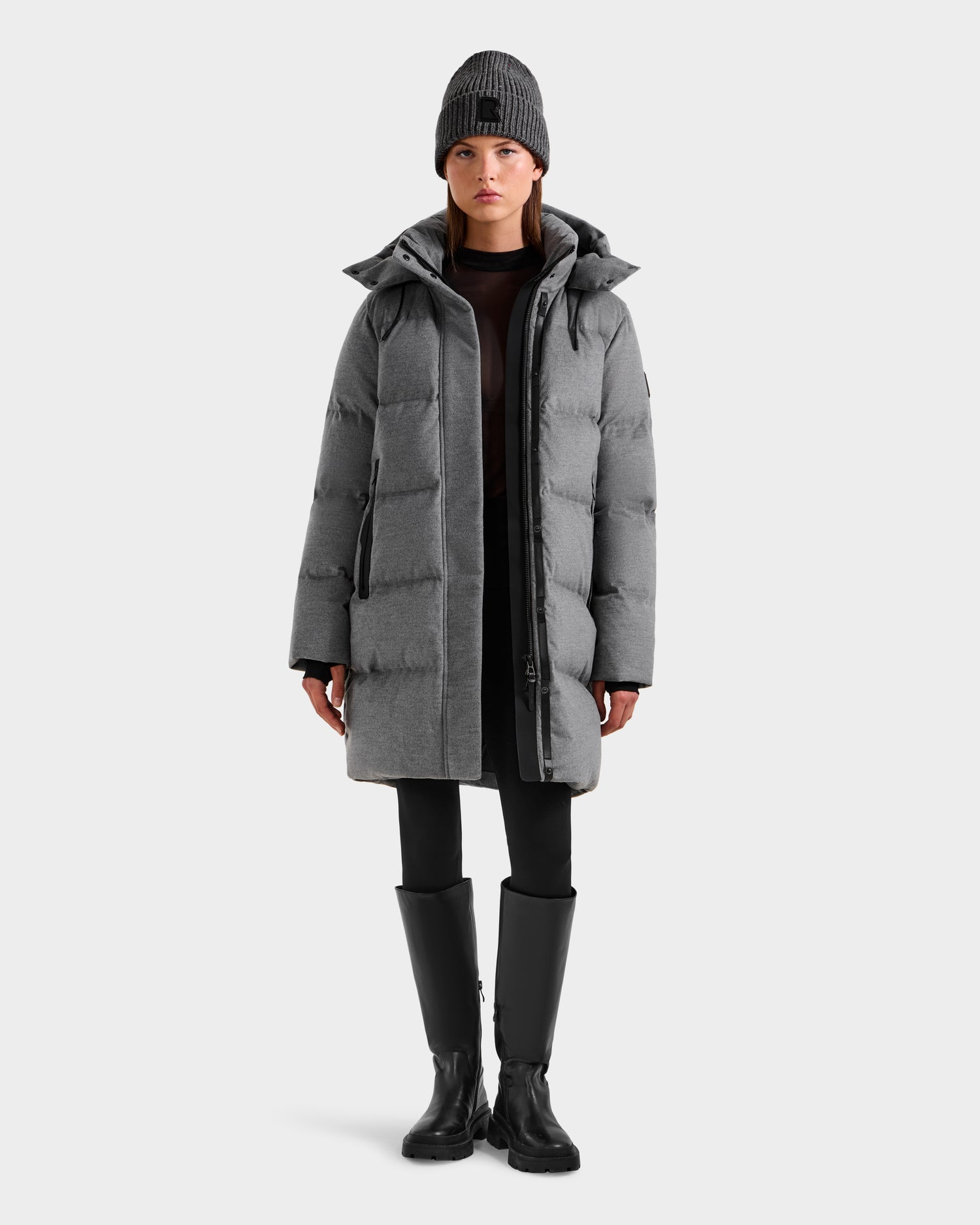 A person faces forward wearing the Rudsak JOELLE W - 8125567 Heather Charcoal women’s down puffer coat, paired with black leggings, knee-high boots, and a gray knit beanie against a plain light background.