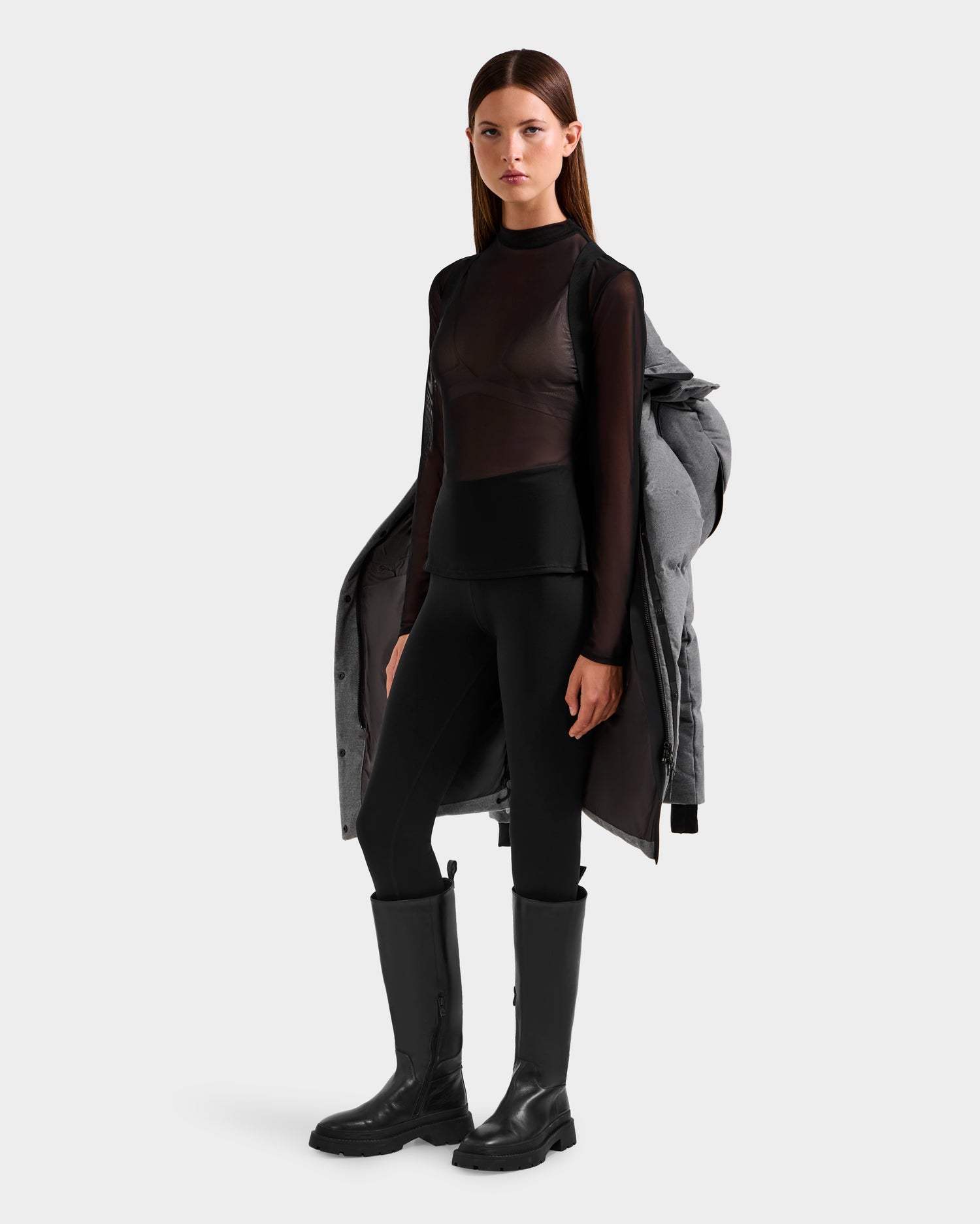 A woman with straight hair and a neutral expression stands facing forward, draping the Rudsak JOELLE W - 8125567 Heather Charcoal wool-blend shell coat over her shoulders. She's wearing a sheer black top, black leggings, and tall black boots.