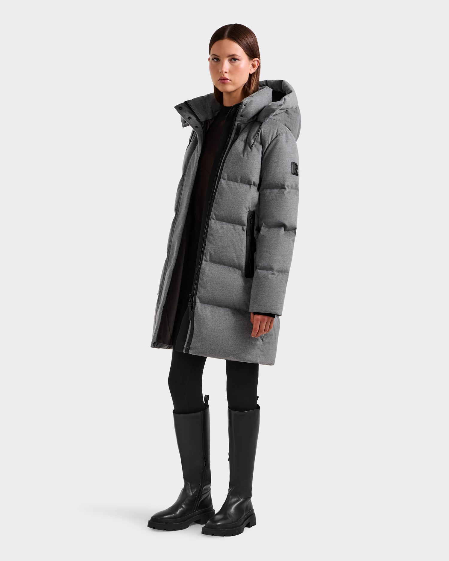 A woman stands facing forward, wearing the Rudsak JOELLE W - 8125567 Heather Charcoal down puffer with a hood, black leggings, and knee-high black boots against a plain light background.