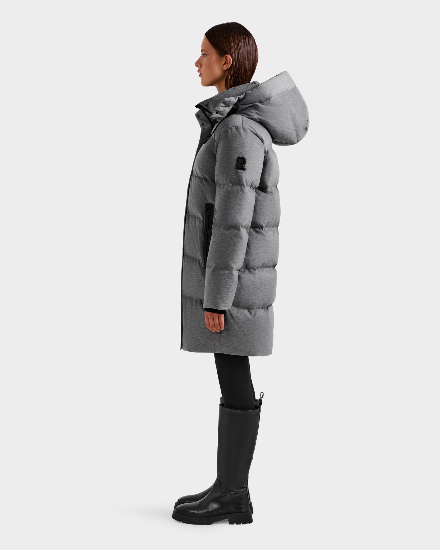 A woman stands in profile wearing the Rudsak JOELLE W - 8125567 Heather Charcoal down puffer with a hood, paired with black leggings and tall boots—a cold-weather essential—set against a plain white background.