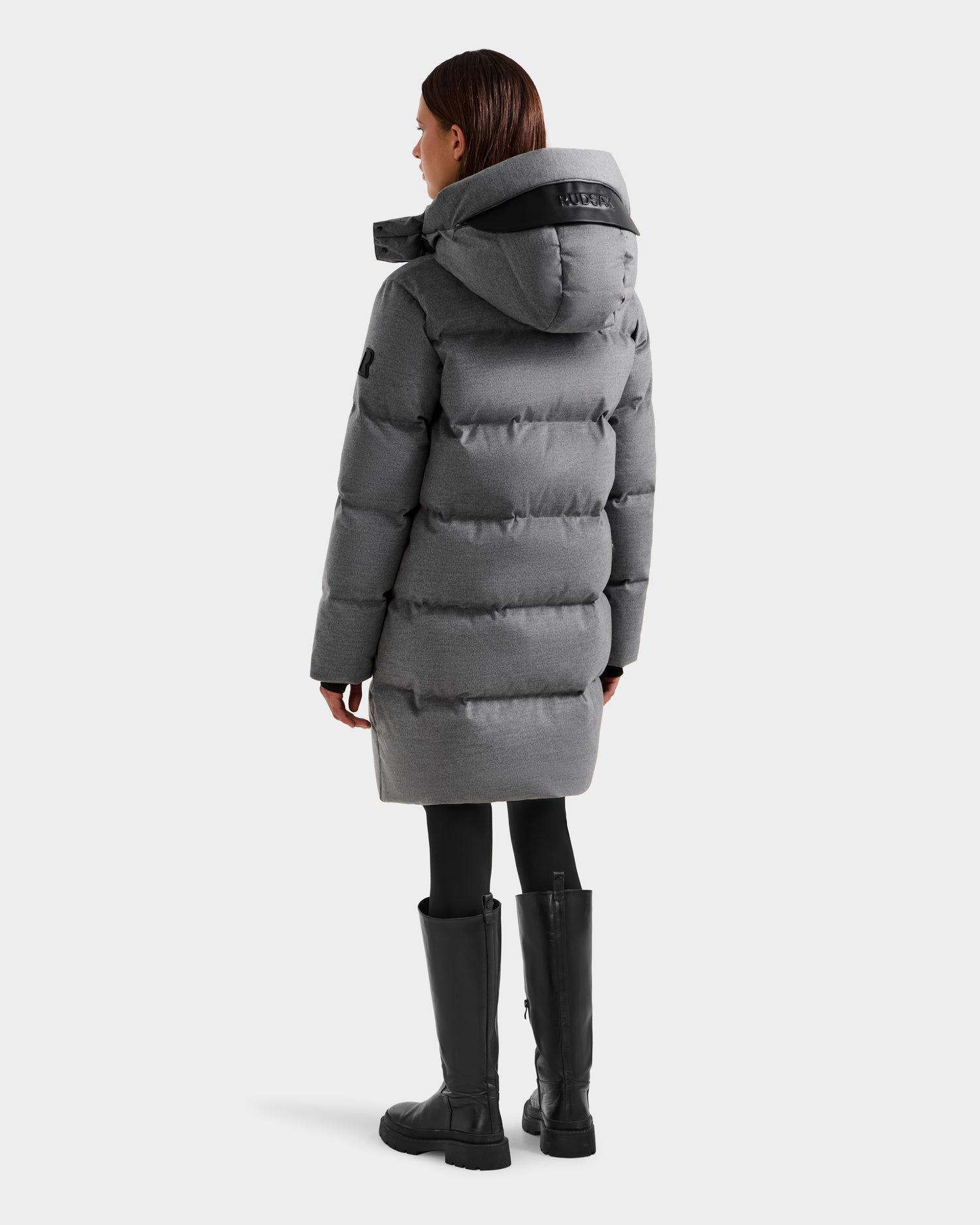 A person wearing the Rudsak JOELLE W - 8125567 Heather Charcoal down puffer coat with hood, black leggings, and tall boots stands facing away from the camera against a plain white background—a perfect cold-weather essential.