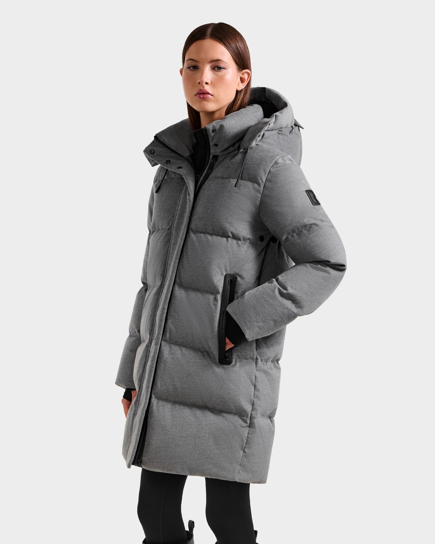 A woman models the Rudsak JOELLE W - 8125567 Heather Charcoal, a long grey down puffer coat with a hood and black zipper details, styled with black pants against a plain light background.