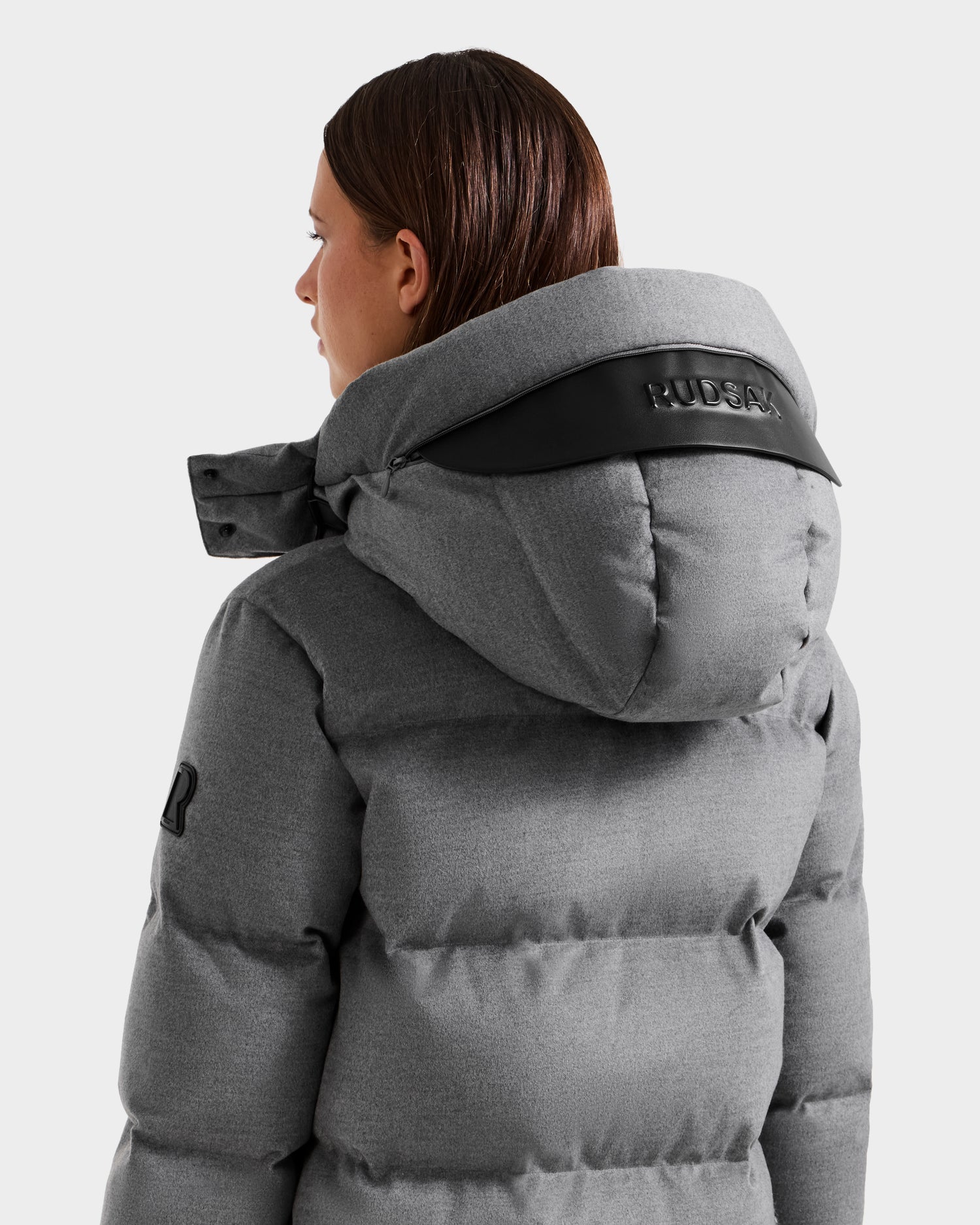 A person is seen from the back wearing the Rudsak JOELLE W - 8125567 puffer in heather charcoal, featuring a large hood embossed with "RUDSAK," black trim, visible stitching, and a sleeve patch—an essential for cold weather.