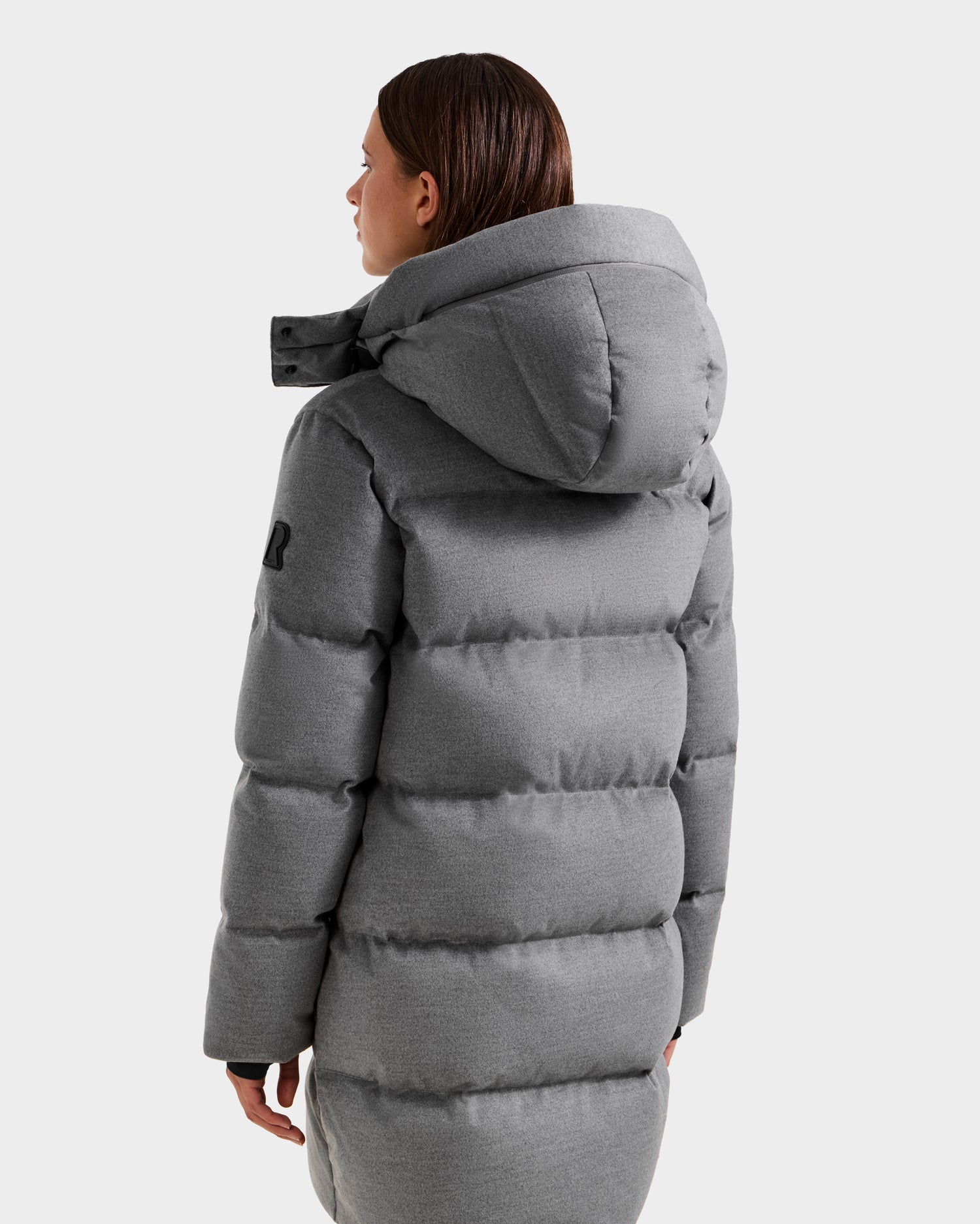 A person with straight brown hair, seen from the back, is wearing the Rudsak JOELLE W - 8125567 Heather Charcoal women’s down puffer jacket with a large hood and black patch on the left upper sleeve against a plain light background.