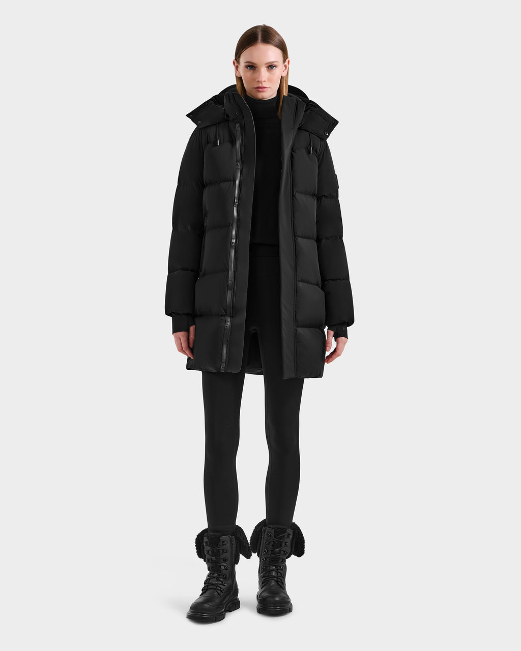 A woman stands facing forward wearing the Rudsak JOELLE - 8124729 Black quilted down coat, paired with a black turtleneck, leggings, and lace-up boots, against a plain white background.