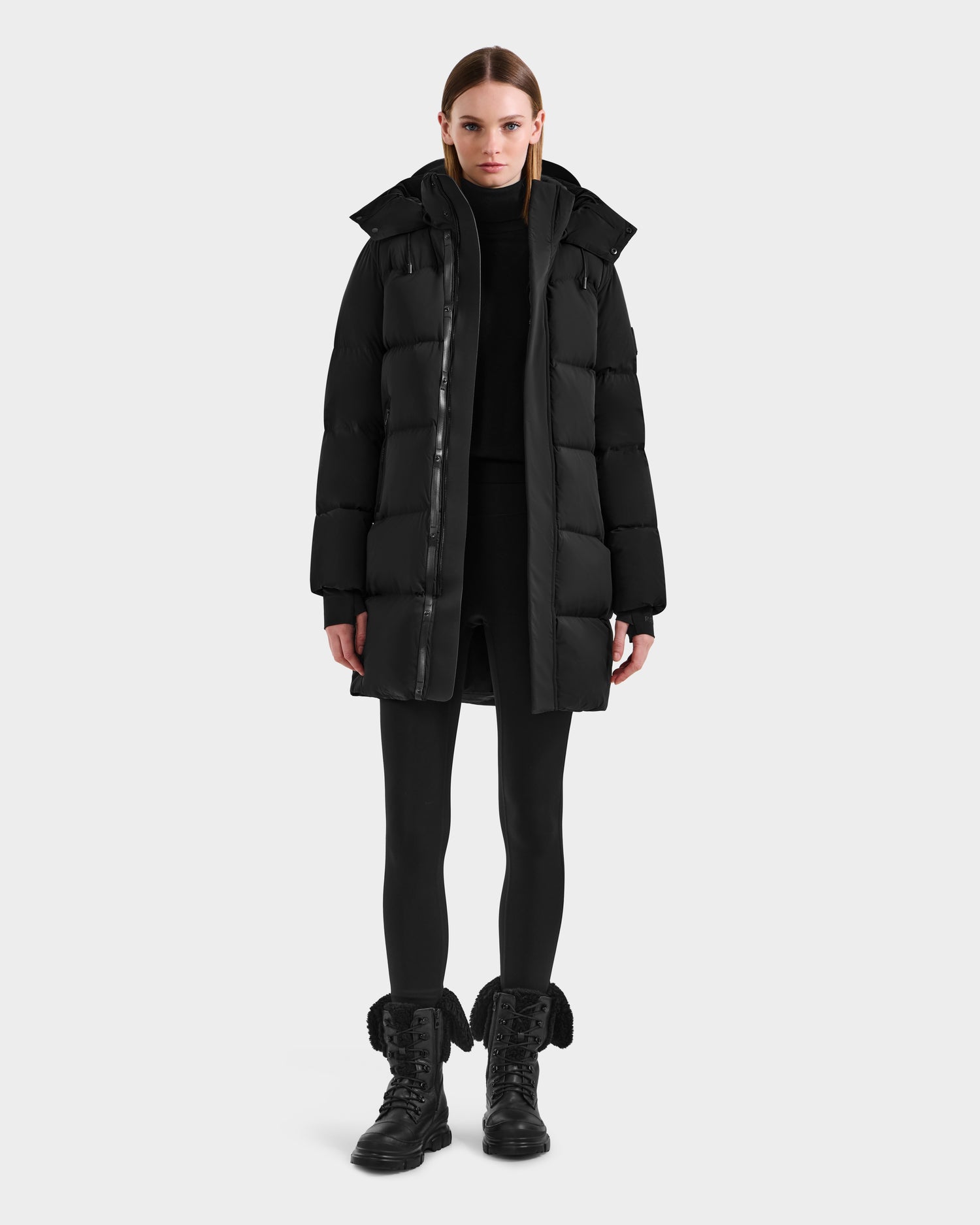 A woman stands facing forward wearing the Rudsak JOELLE - 8124729 Black quilted down coat, paired with a black turtleneck, leggings, and lace-up boots, against a plain white background.