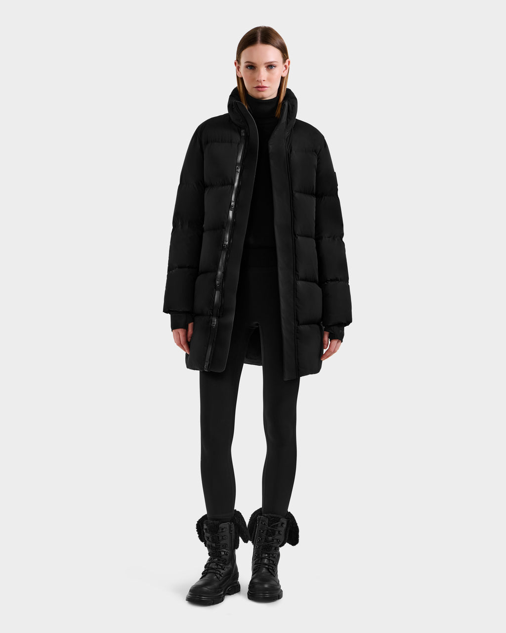 A woman faces forward in Rudsak’s JOELLE - 8124729 Black women's quilted down coat with RDS-certified down, paired with black leggings, a black turtleneck, and black lace-up boots against a plain light gray background.