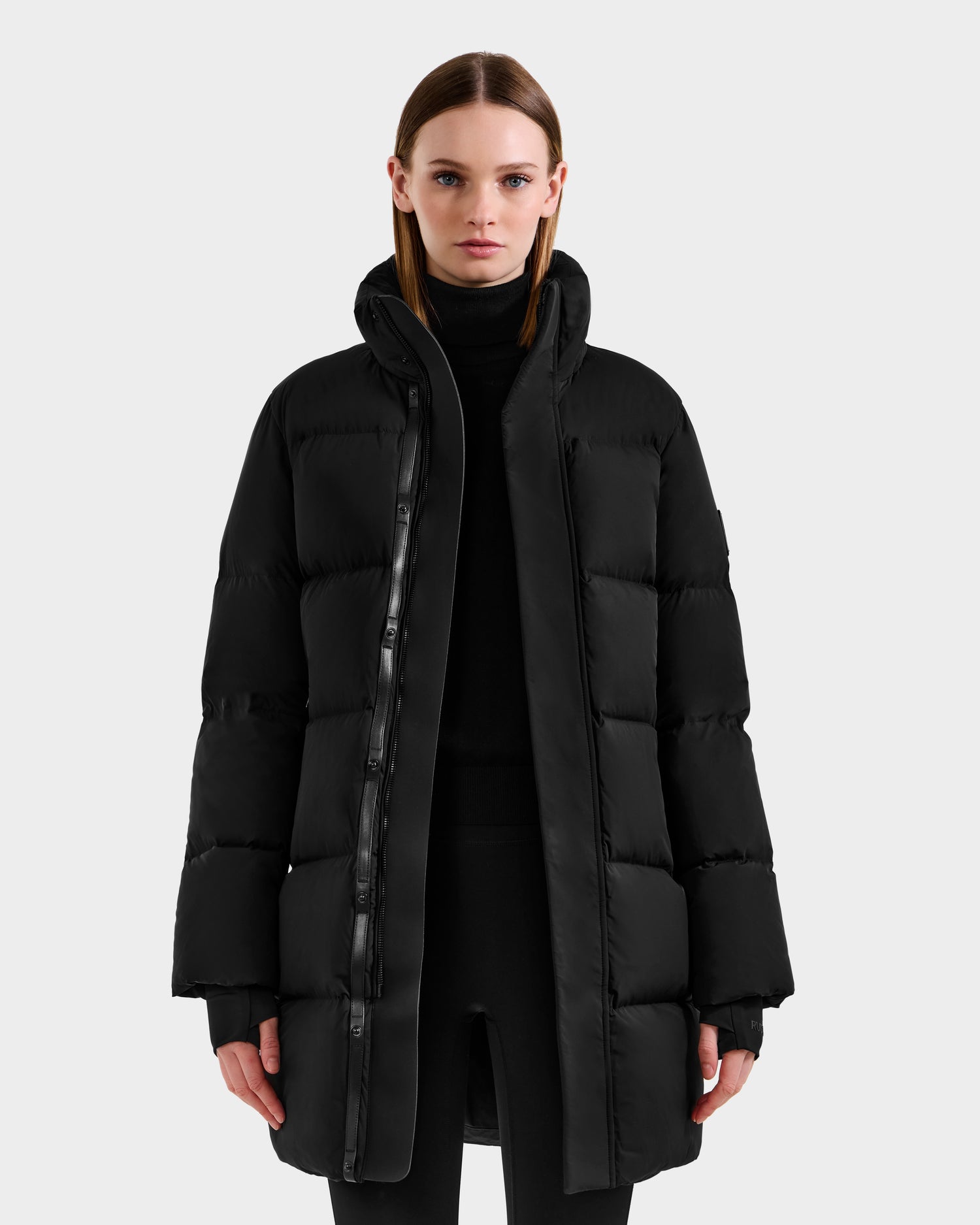 A woman faces forward, wearing the Rudsak JOELLE - 8124729 Black women’s quilted down coat with a high collar over black clothing. The RDS-certified winter coat stands out against a plain, light gray background.