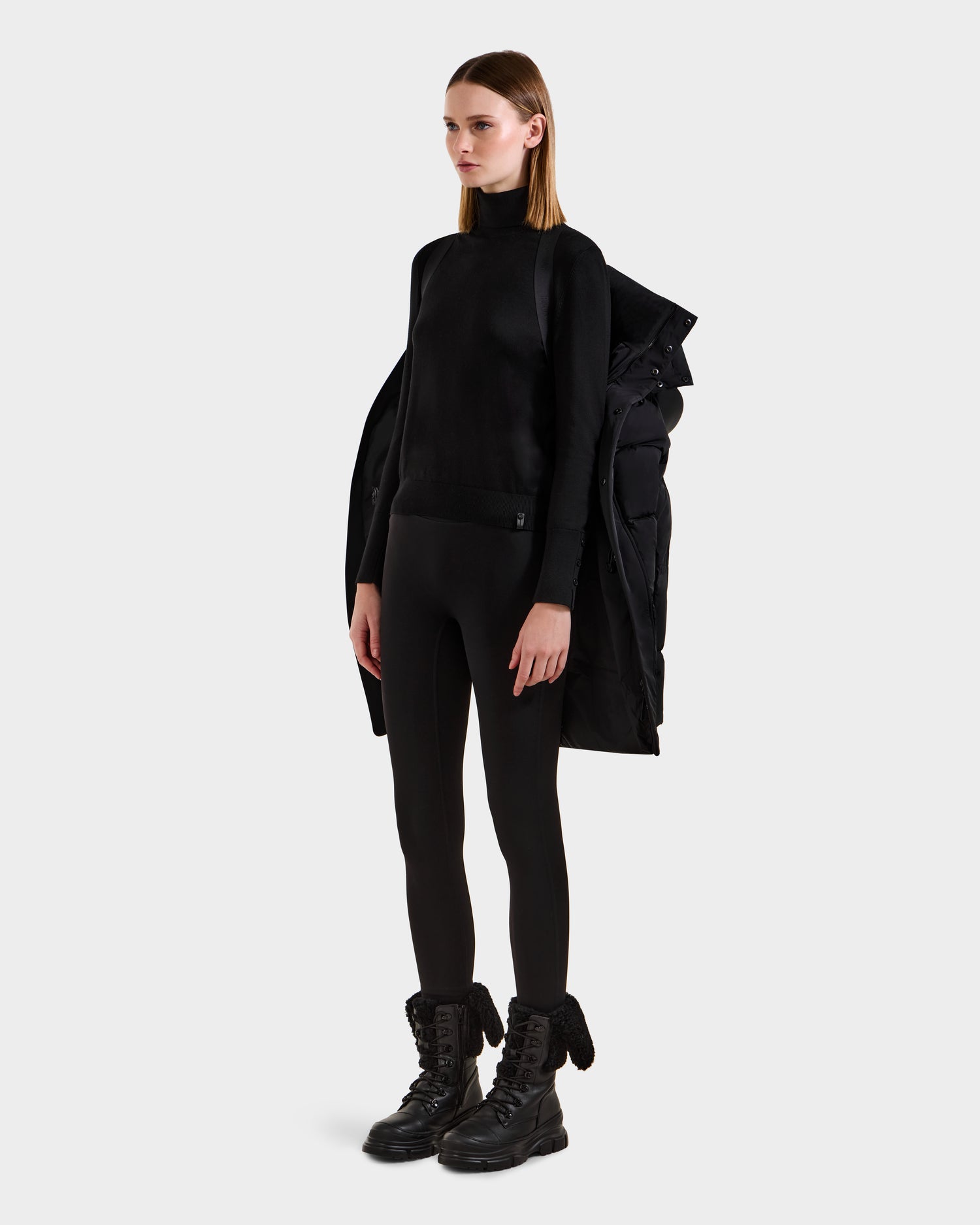 A woman poses against a plain background in a black turtleneck, black leggings, chunky lace-up boots, and the Rudsak JOELLE - 8124729 Black women's quilted down coat draped over her shoulders.