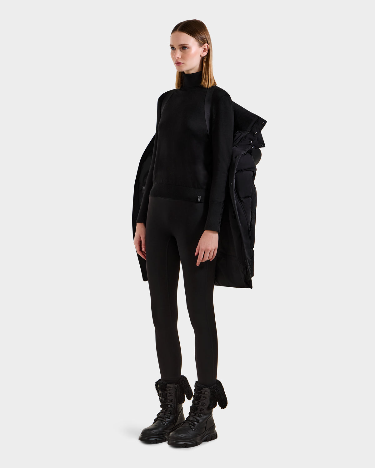 A woman poses against a plain background in a black turtleneck, black leggings, chunky lace-up boots, and the Rudsak JOELLE - 8124729 Black women's quilted down coat draped over her shoulders.