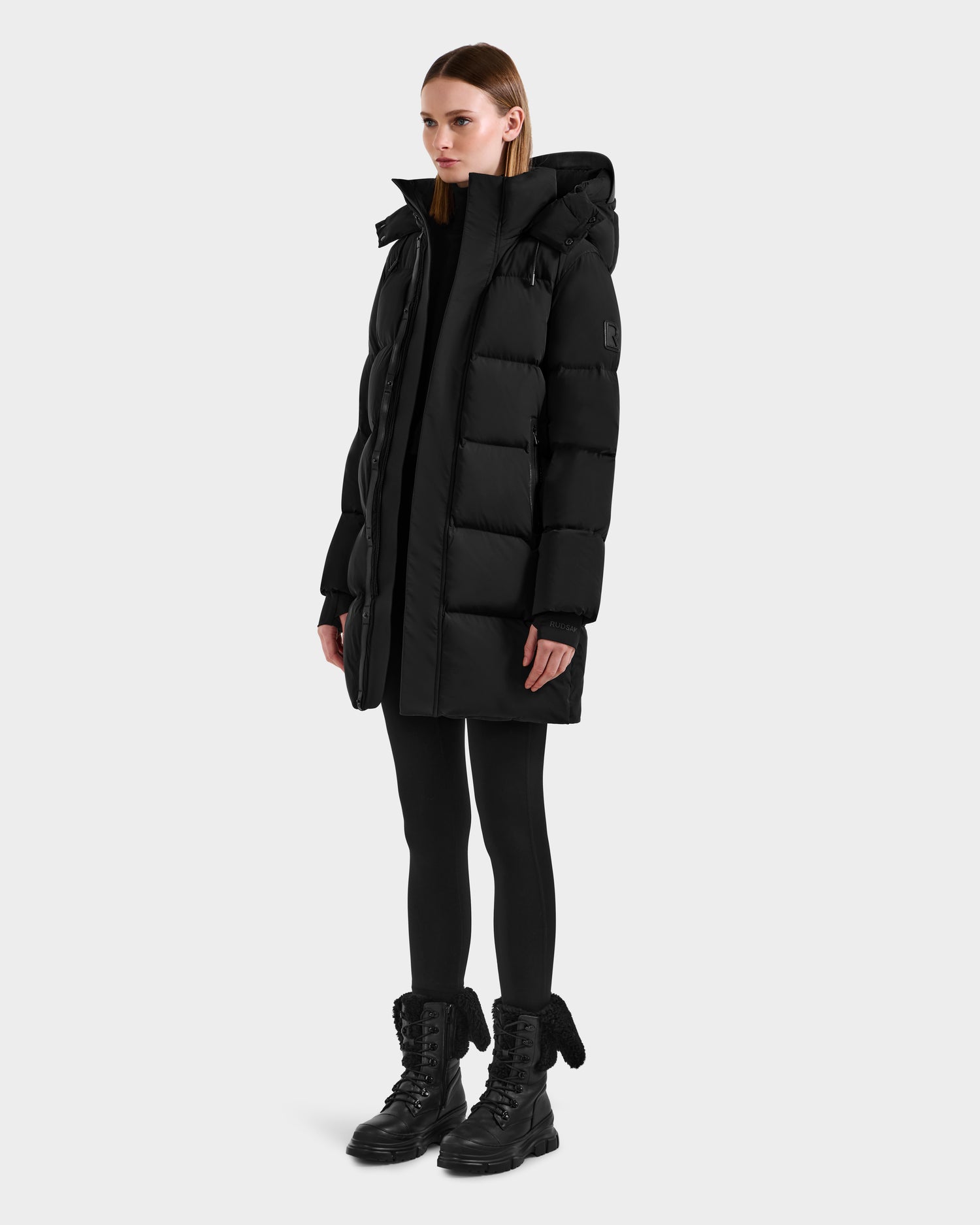A person wears the Rudsak JOELLE - 8124729 Black women's quilted down coat with a hood, styled with black leggings and lace-up boots, set against a plain light gray background.