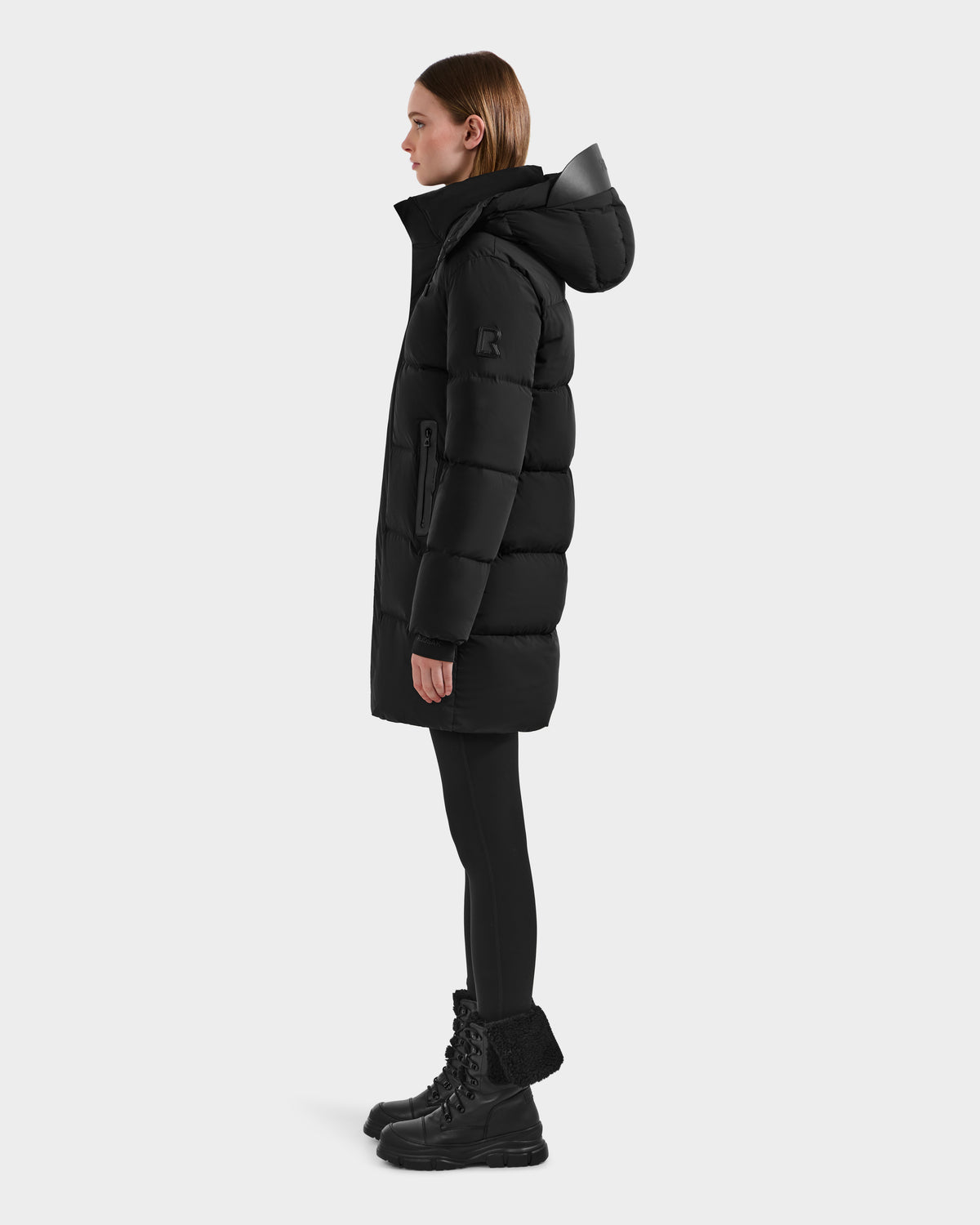 A woman stands in profile wearing the Rudsak JOELLE - 8124729 Black quilted down coat, paired with black leggings and black lace-up boots, against a plain white background.
