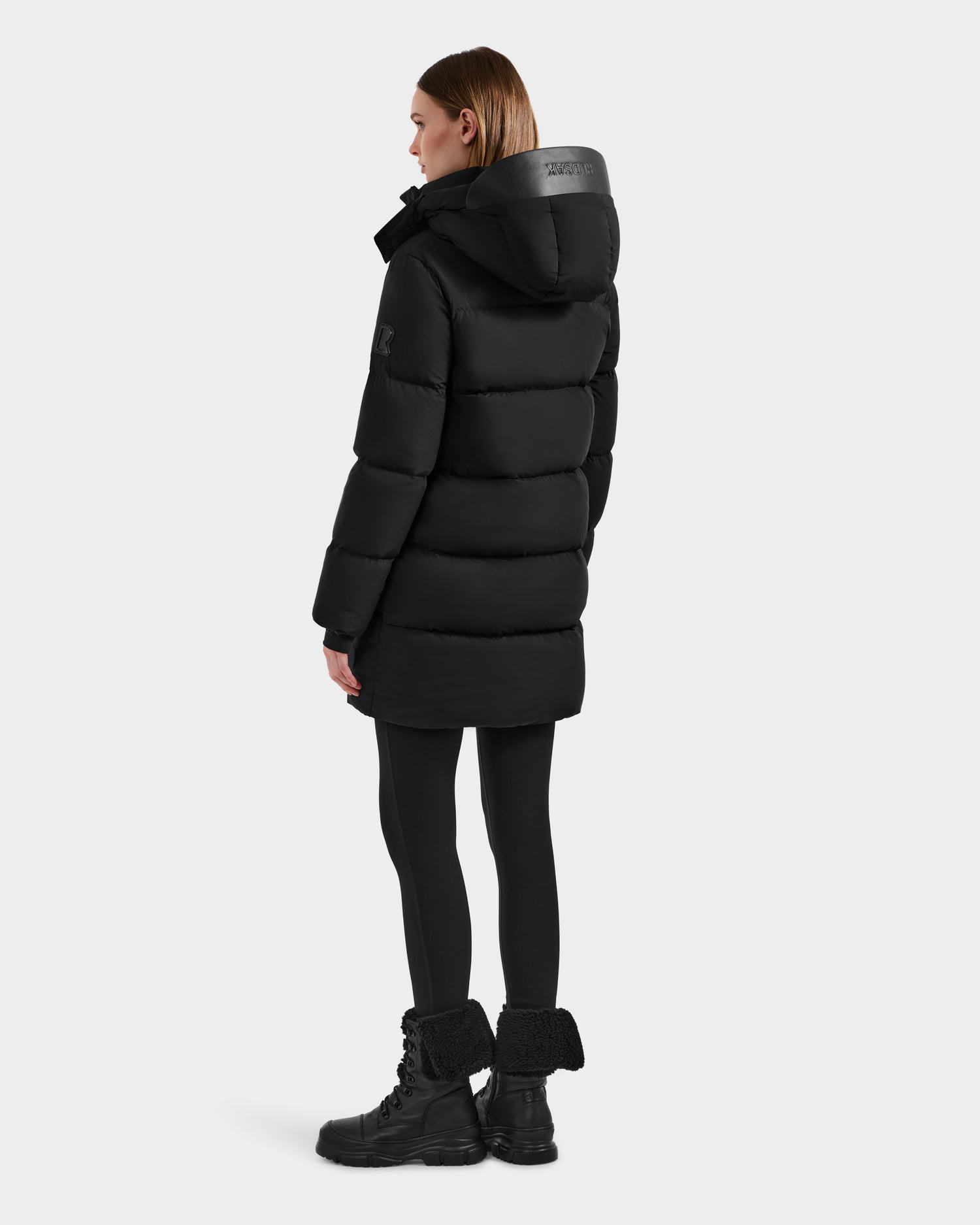 A woman faces away on a light background, wearing the Rudsak JOELLE - 8124729 Black women's quilted down coat with a hood, paired with black leggings and black lace-up boots.