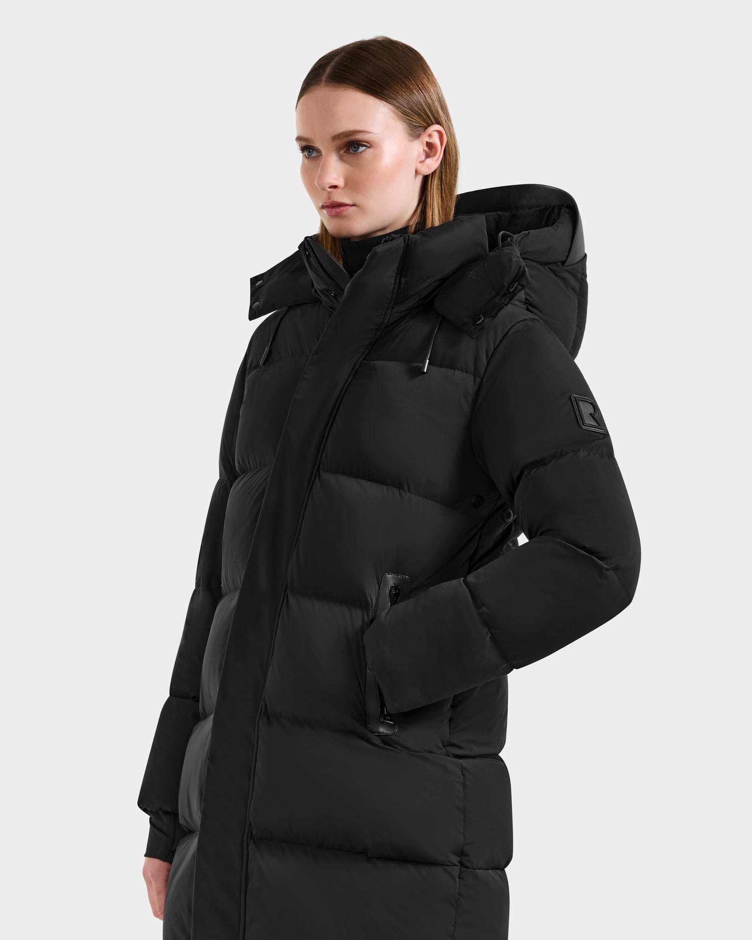 A person wearing the Rudsak JOELLE - 8124729 Black women's quilted down coat with a hood, standing against a plain light background and looking to the side—a timeless winter essential.