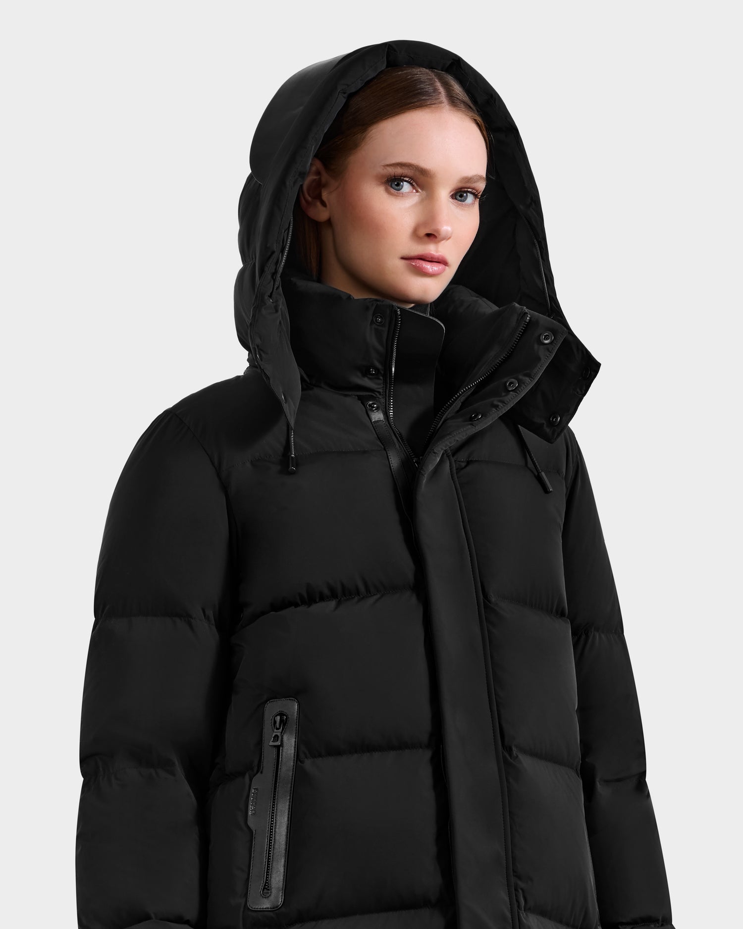 A woman stands against a light gray background wearing the Rudsak JOELLE - 8124729 Black quilted down coat with a hood, filled with RDS-certified down. She has straight brown hair and looks slightly to the side with a neutral expression.