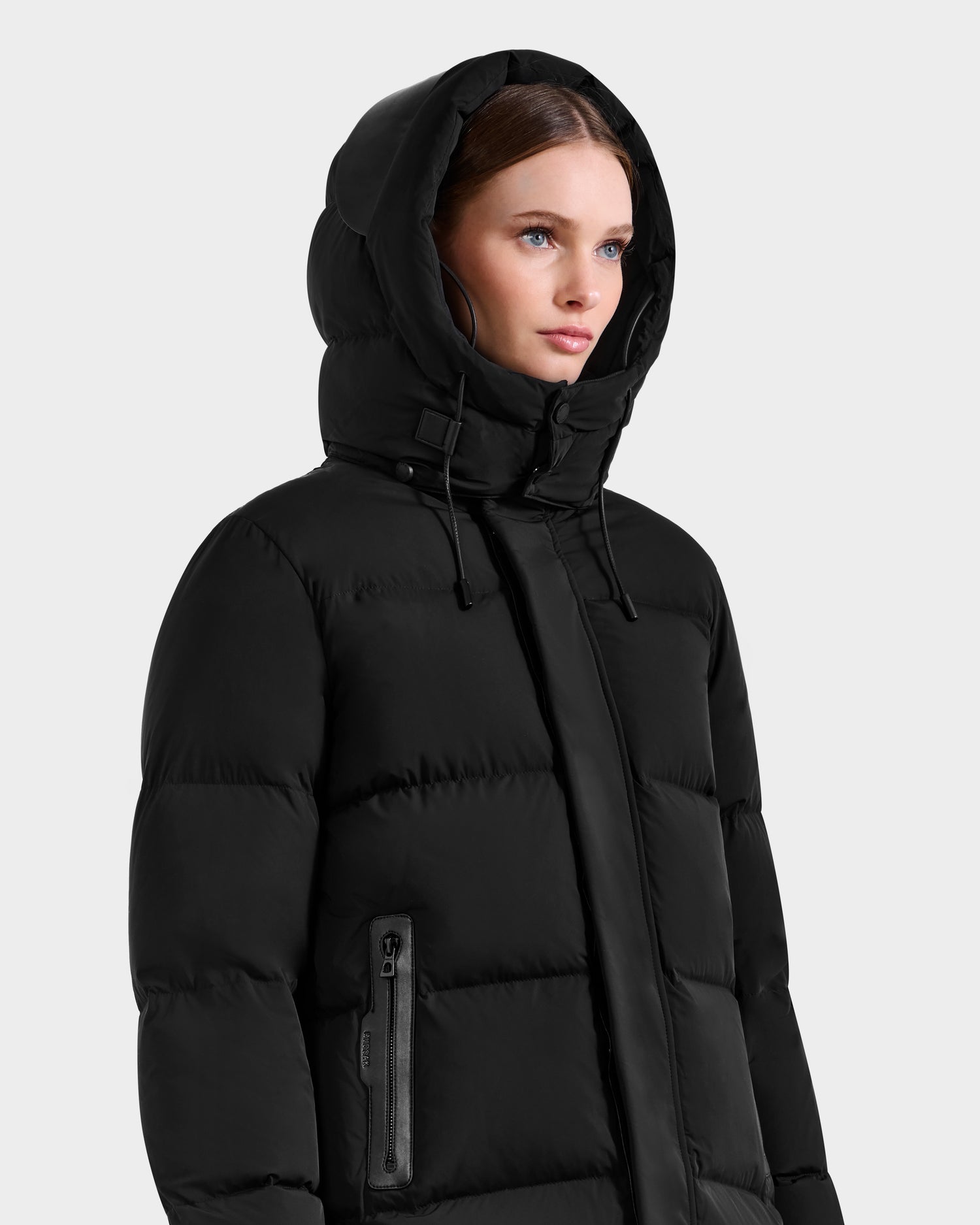 A woman models the Rudsak JOELLE - 8124729 Black quilted down coat, featuring a high collar, zipper accents, and hood drawstrings, against a light grey background.