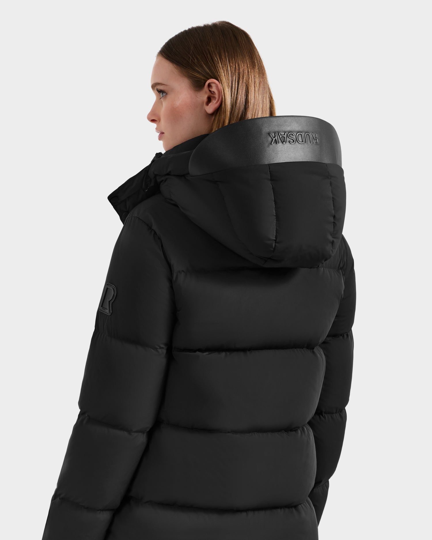 A woman with brown hair wears the JOELLE - 8124729 Black quilted down coat with a hood by Rudsak. She stands slightly turned to the side against a plain light gray background.