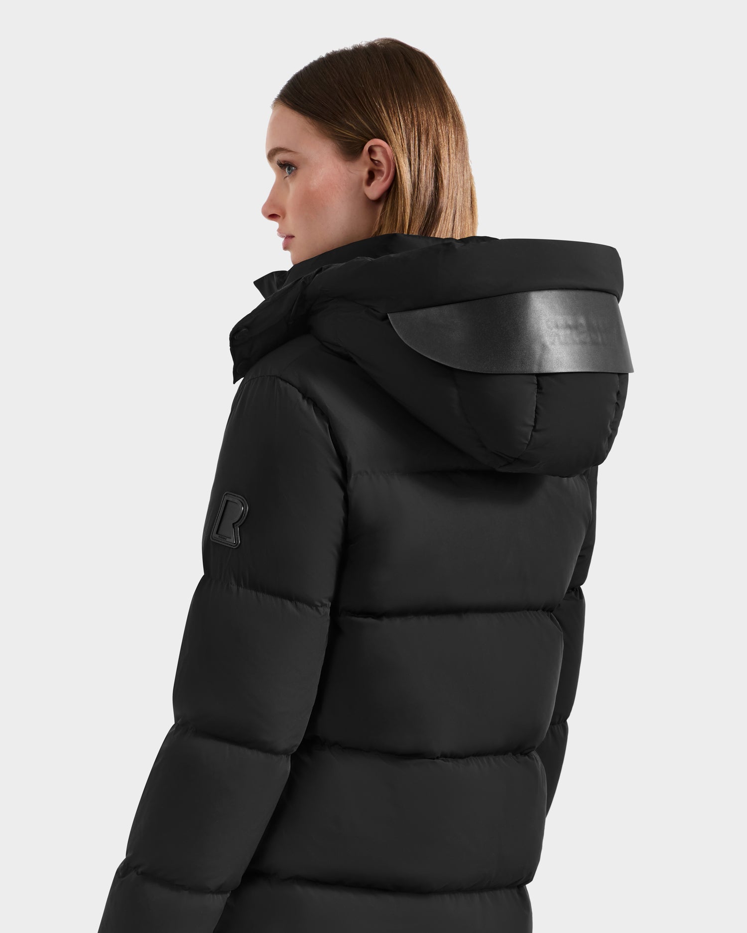 A woman with straight, shoulder-length brown hair models the Rudsak JOELLE - 8124729 Black women's quilted down coat with a hood and leather-like accents, shown from the back and in profile against a plain light background.