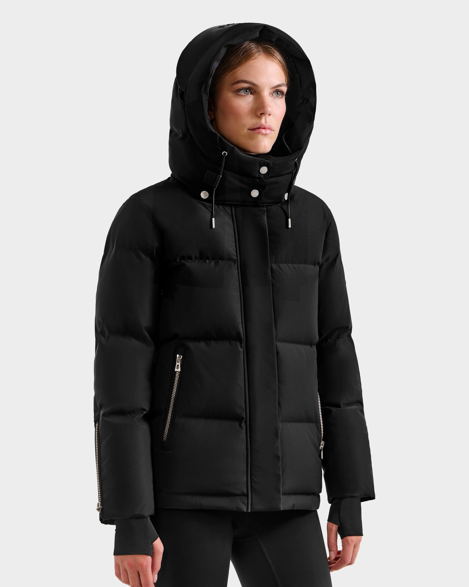 A woman wears the Rudsak JOON H - 8125586 Black hooded, insulated winter jacket with zippered side pockets and fitted cuffs, standing against a plain light gray background.