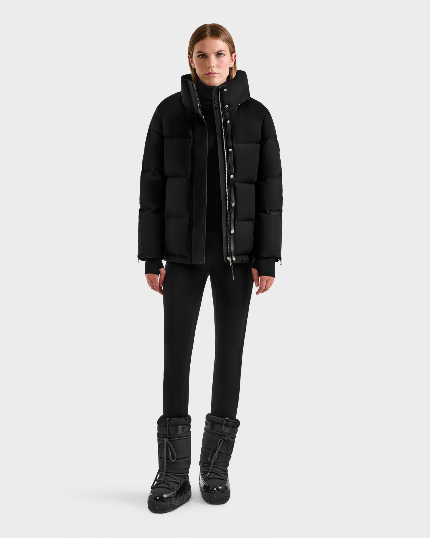 A woman stands facing forward against a light gray background, wearing the Rudsak JOON H - 8125586 Black insulated winter jacket, paired with black pants and black boots.