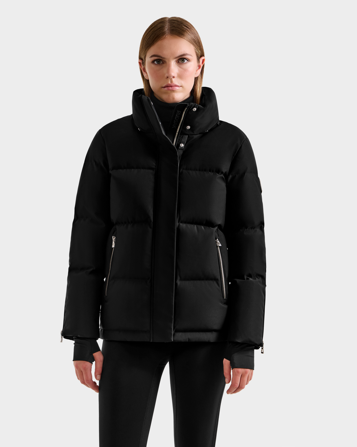 A woman faces forward, wearing the Rudsak JOON H - 8125586 Black insulated winter jacket with a high collar and zippered pockets, paired with black leggings. She has straight, shoulder-length hair against a plain light gray background.