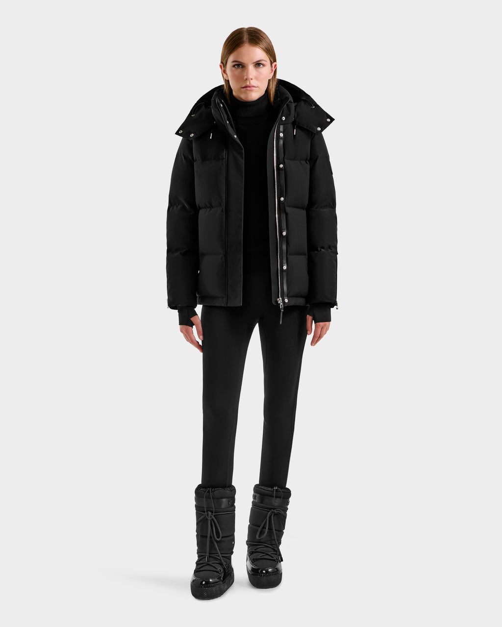 A woman faces forward, wearing the Rudsak JOON H - 8125586 Black insulated winter jacket, paired with a black turtleneck, pants, and boots against a plain light gray background.