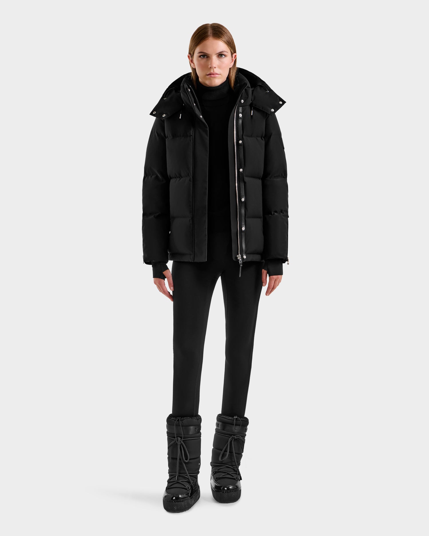 A woman faces forward, wearing the Rudsak JOON H - 8125586 Black insulated winter jacket, paired with a black turtleneck, pants, and boots against a plain light gray background.