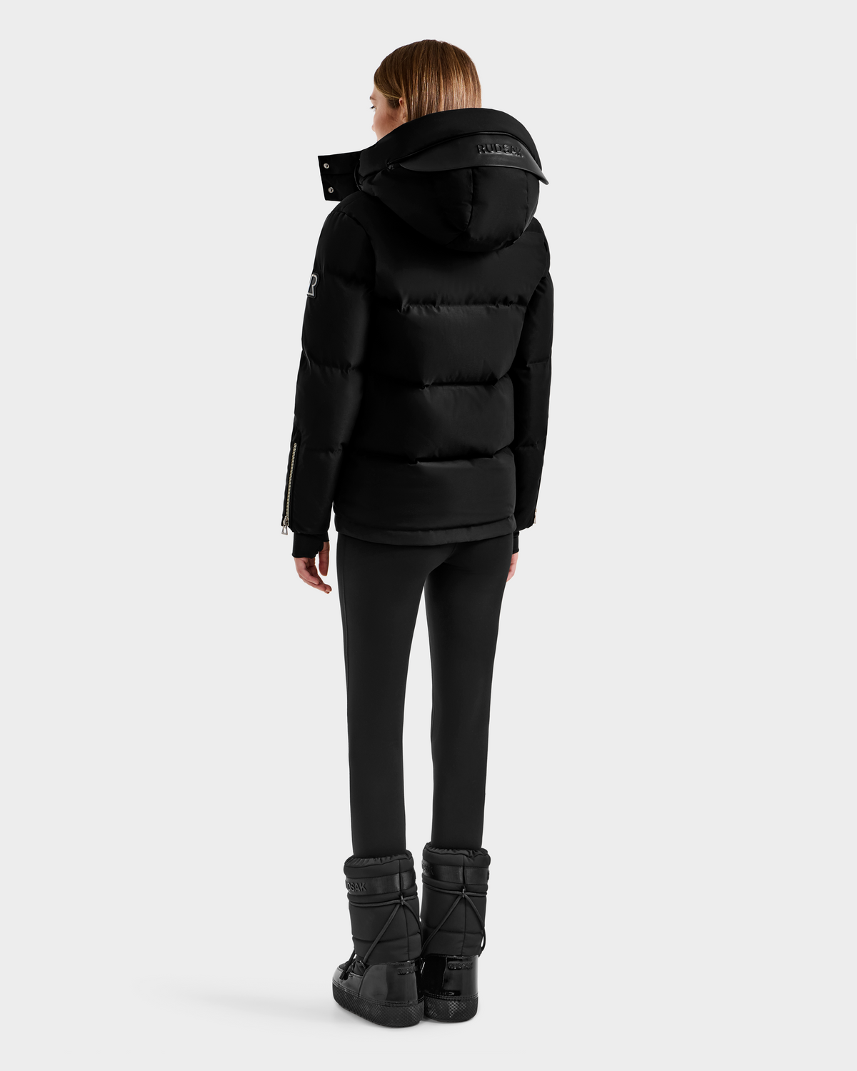A person faces away in the Rudsak JOON H - 8125586 Black women’s down jacket with a hood, paired with black leggings and boots against a light gray background—a must-have cold-weather piece.