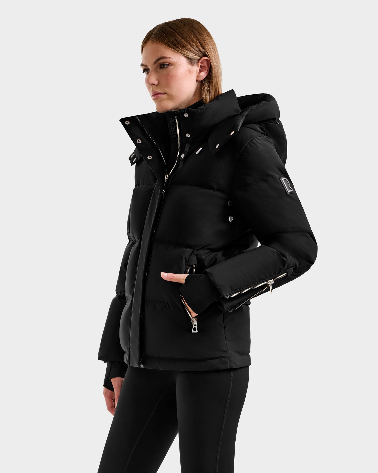 A woman wears the Rudsak JOON H - 8125586 Black RDS-certified down winter coat with a hood, featuring silver zippers and snap buttons, as she stands against a plain light background with her left hand in the pocket.