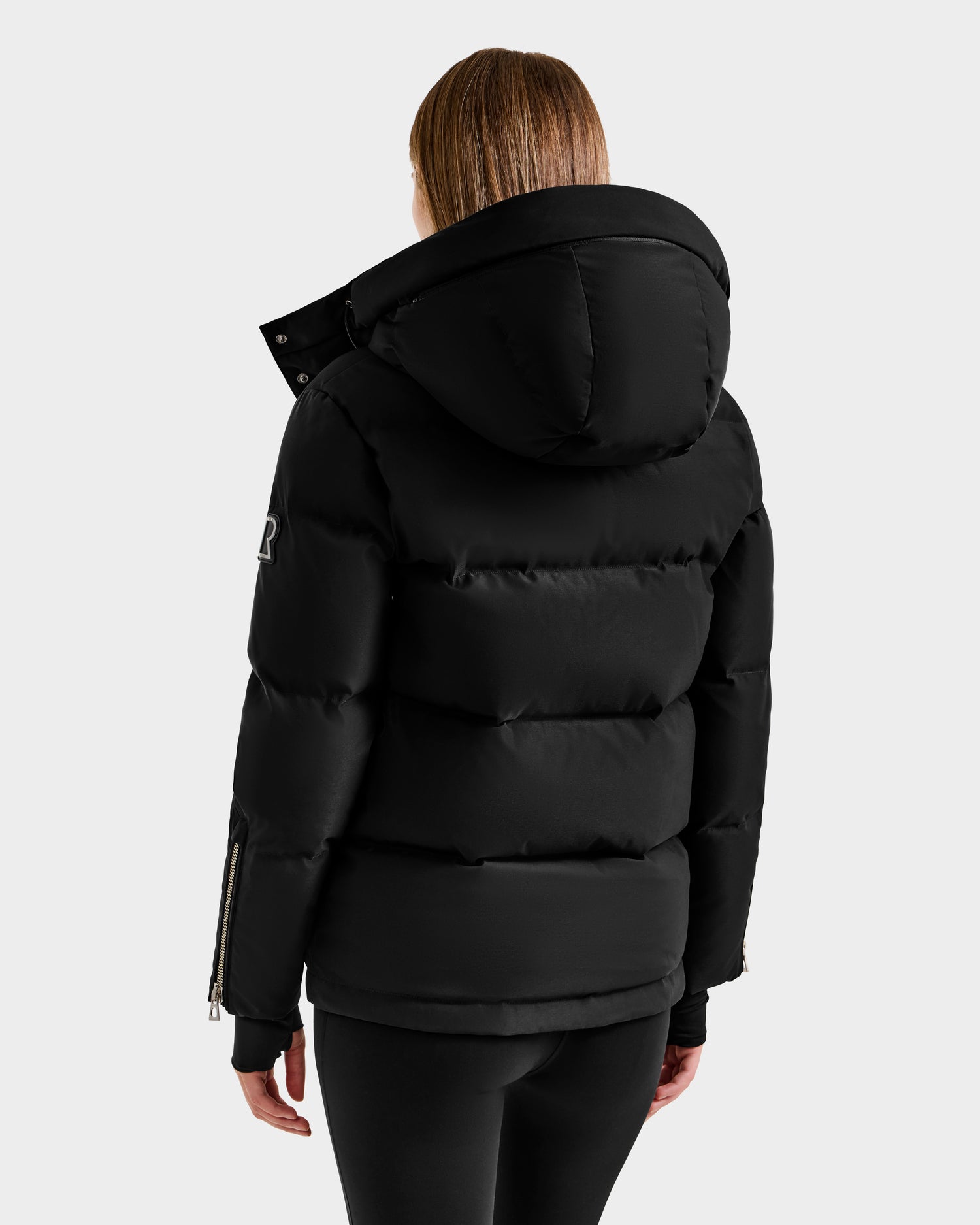 A person with straight light brown hair, seen from behind, wears the Rudsak JOON H - 8125586 Black insulated winter jacket with a large hood and zippered sleeves, paired with black leggings, against a plain light backdrop.