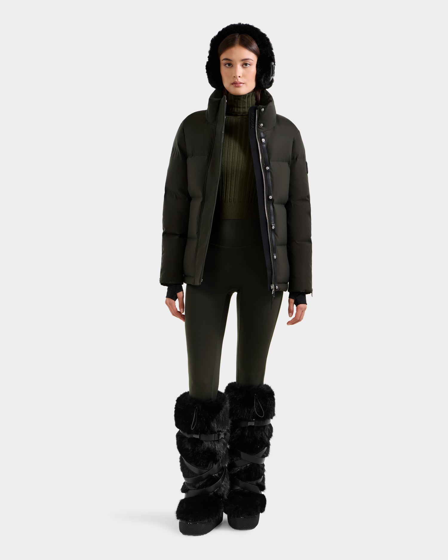 A woman stands facing forward, wearing the Rudsak JOON H - 8125586 Dark Olive women’s down jacket with a hood, an olive green sweater, matching leggings, and tall black furry boots against a plain white background.
