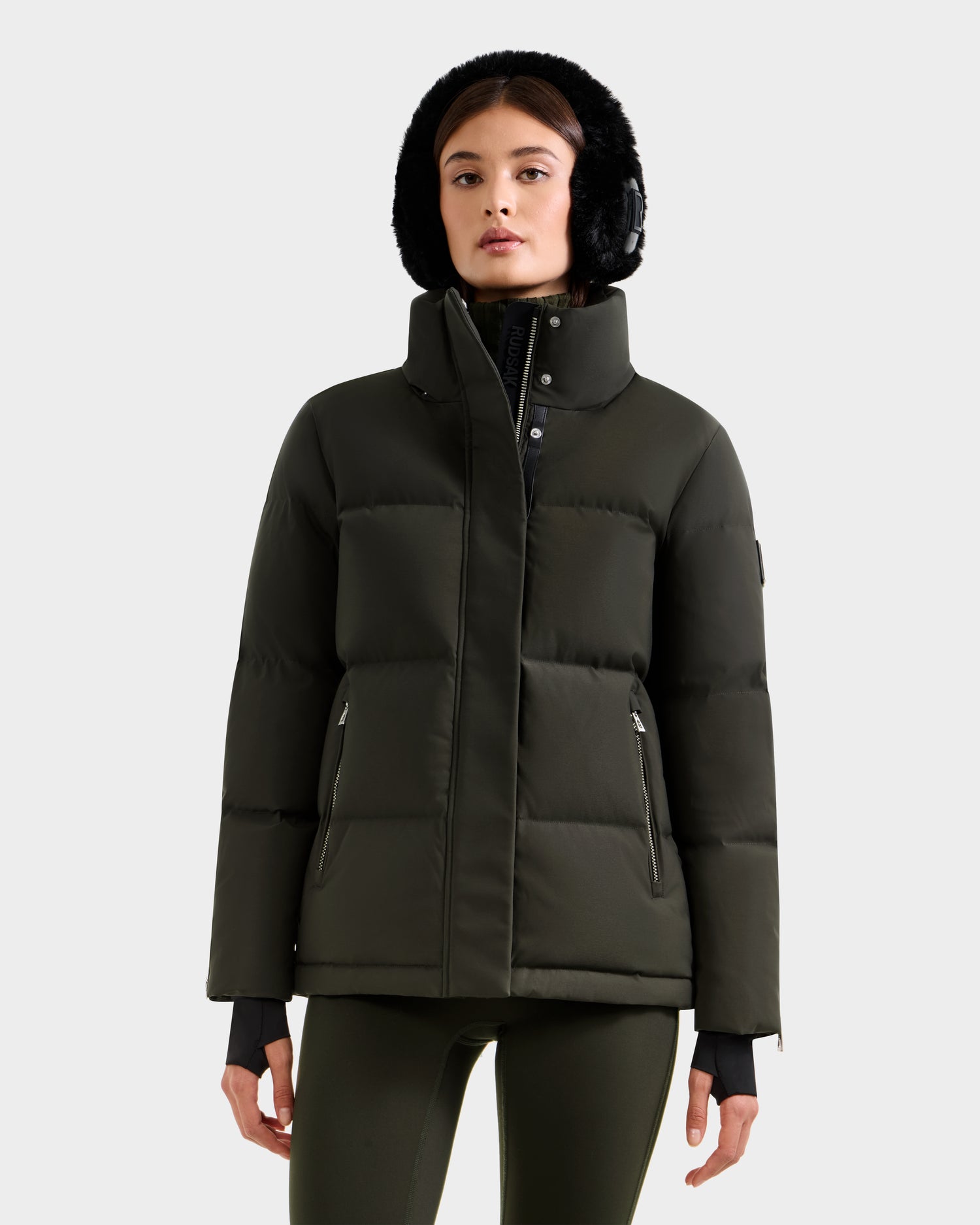 A woman faces forward wearing the Rudsak JOON H - 8125586 Dark Olive insulated jacket with a high collar, zippered pockets, and a black fur-lined hood. She pairs it with matching leggings against a plain light gray background.