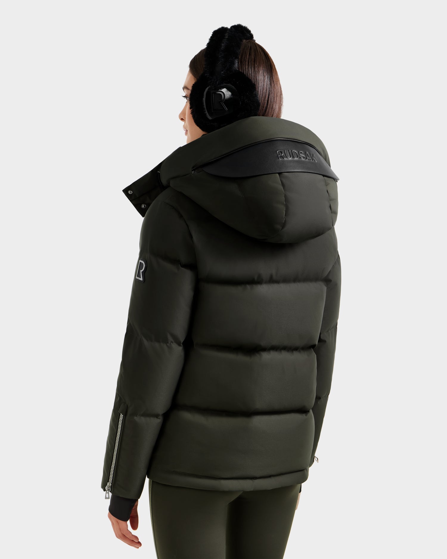 A person seen from the back wears the Rudsak JOON H - 8125586 Dark Olive women’s down jacket with a large hood and logo on the shoulder, paired with black earmuffs, set against a plain light gray background.