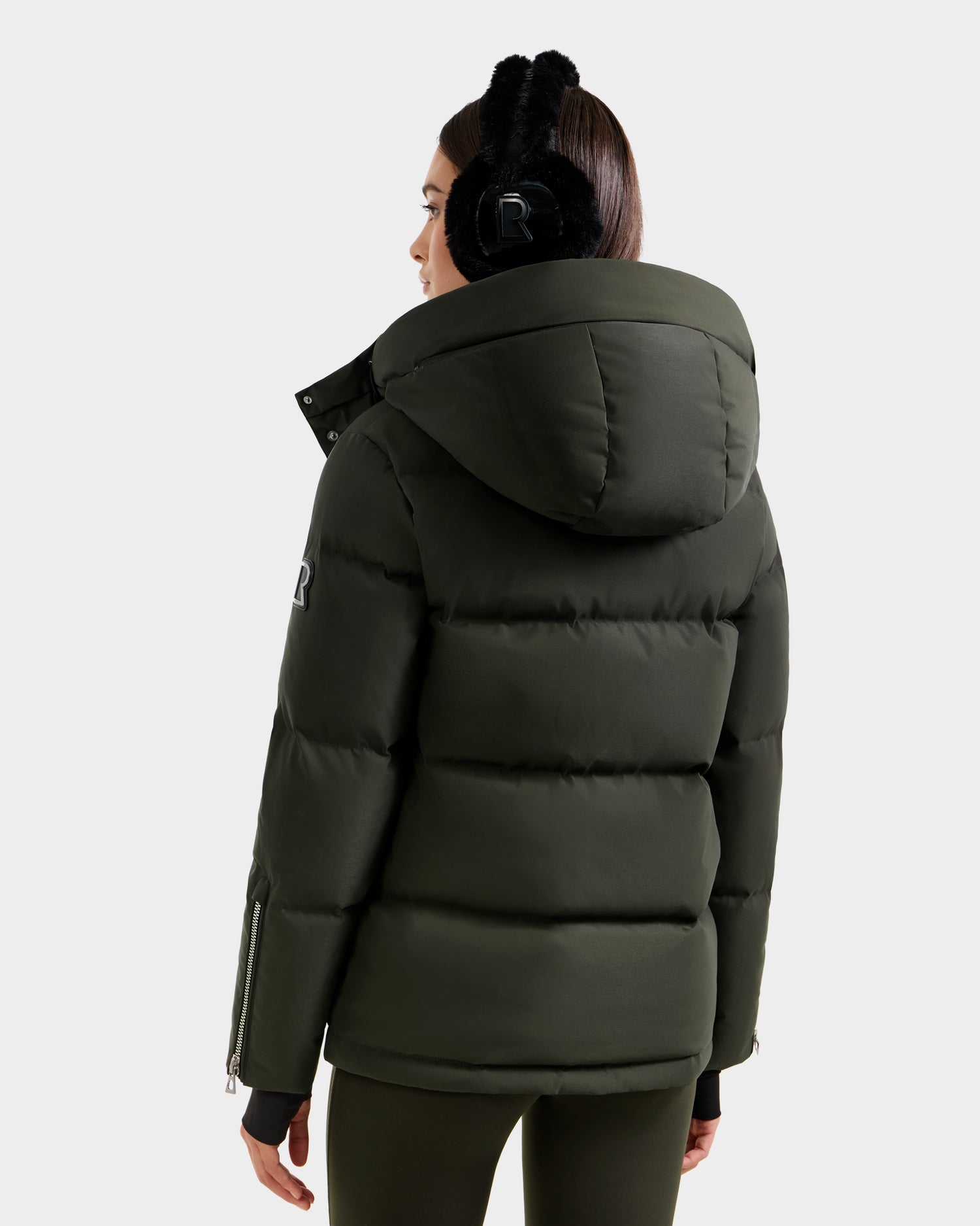 A woman in the Rudsak JOON H - 8125586 Dark Olive puffer jacket, featuring a large hood, and black earmuffs stands with her back to the camera against a plain light background.