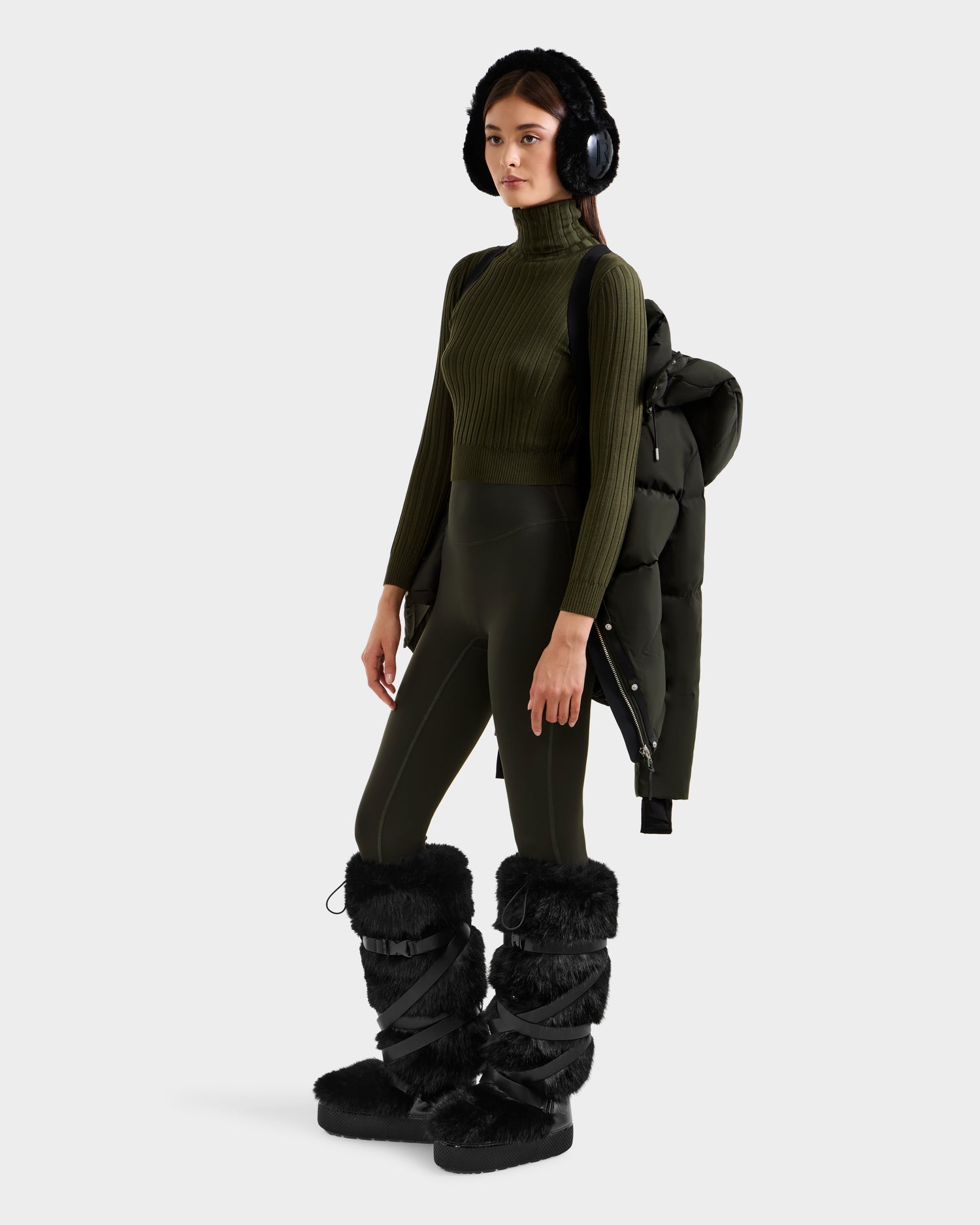 Wearing the Rudsak JOON H - 8125586 Dark Olive ribbed turtleneck and matching leggings, she pairs black furry earmuffs, snow boots, and a jacket over one shoulder—an essential cold-weather look against a white background.