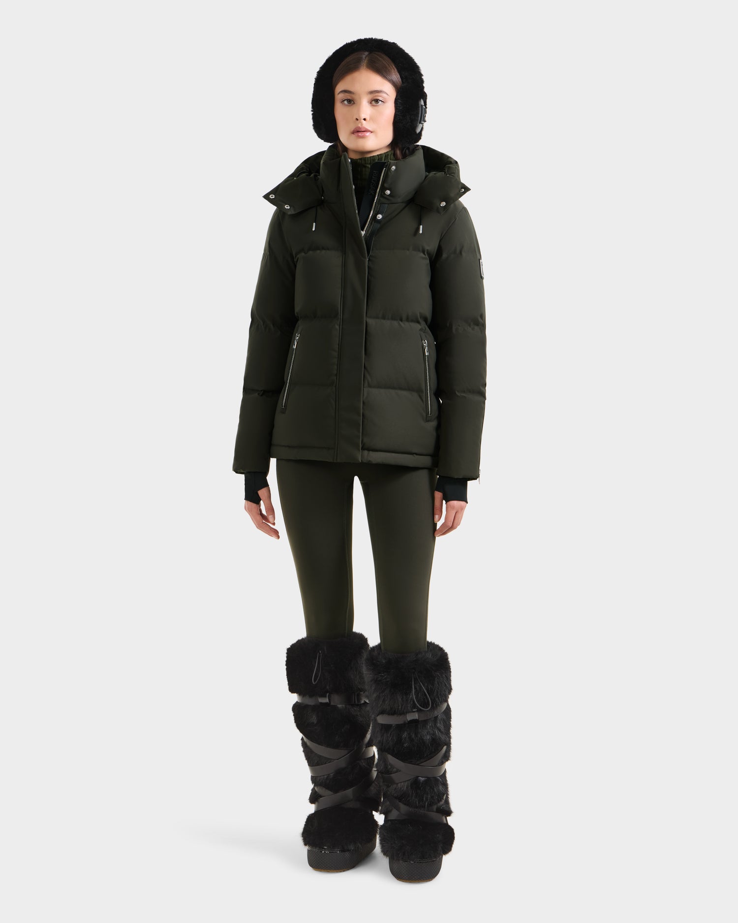A person faces forward against a plain white background, wearing the Rudsak JOON H - 8125586 Dark Olive quilted jacket with hood, matching green leggings, and tall black fur boots.