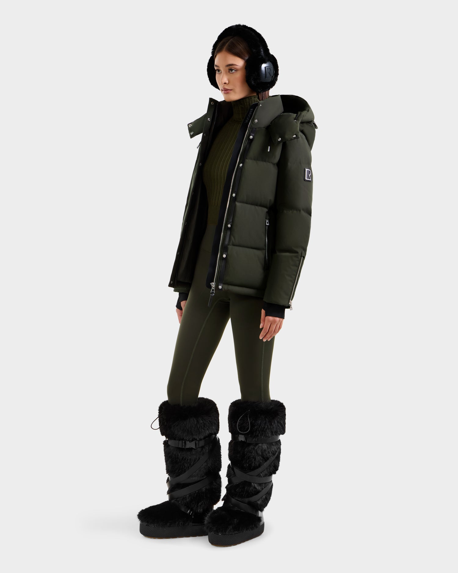 A person wearing black earmuffs, a green women’s down jacket, JOON H - 8125586 Dark Olive leggings by Rudsak, and tall black fur boots stands against a plain light background.