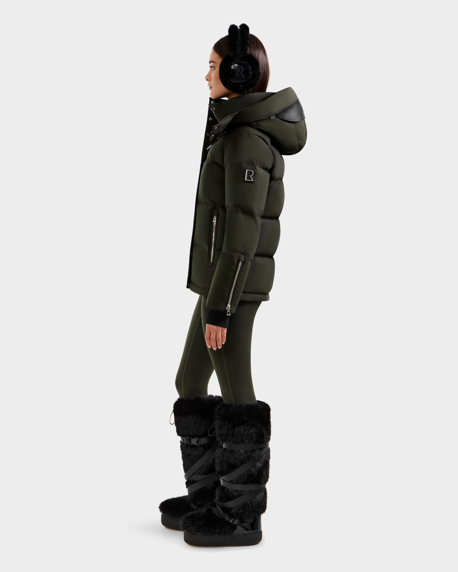 A woman in profile wearing earmuffs, fitted pants, tall black fur boots, and the JOON H - 8125586 Dark Olive women's down jacket by Rudsak stands against a plain light background.