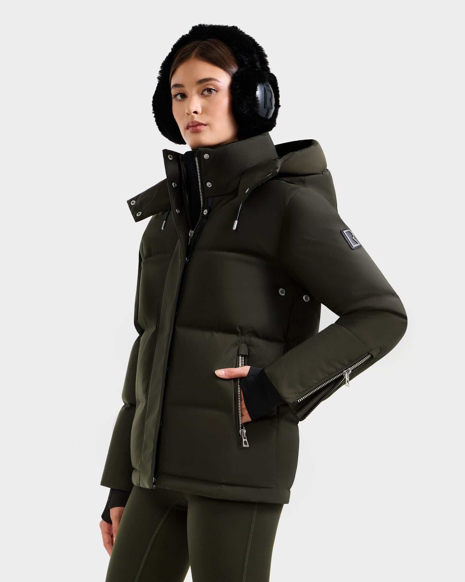 A woman wears the Rudsak JOON H - 8125586 Dark Olive down jacket with a hood, black earmuffs, and matching green pants. She stands against a light background, turned slightly to the side, highlighting the jacket’s lightweight warmth.
