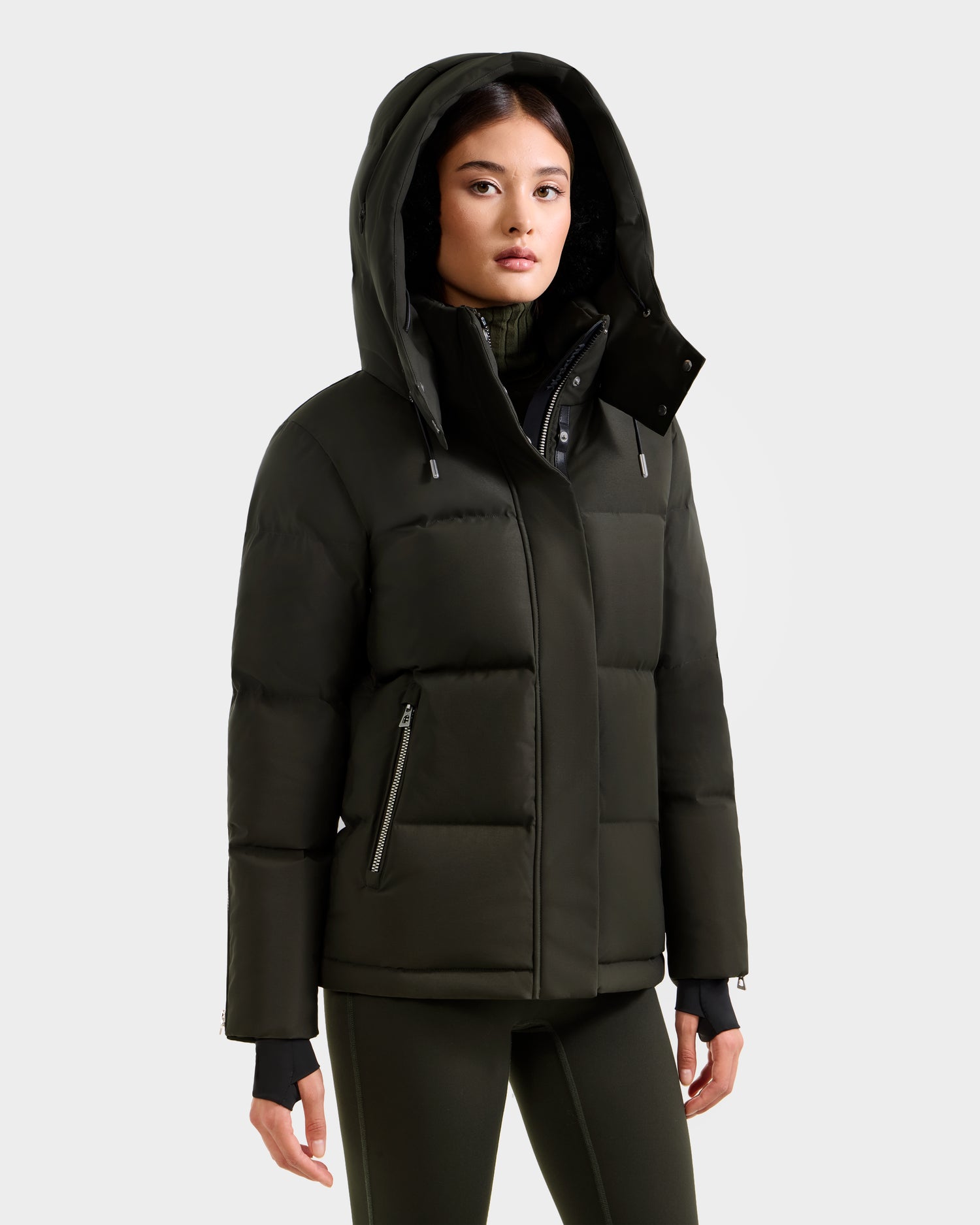 A woman wears the Rudsak JOON H - 8125586 Dark Olive women’s down jacket, featuring zippered pockets. She stands facing forward with her hands at her sides, paired with matching dark green pants for a stylish cold-weather look.