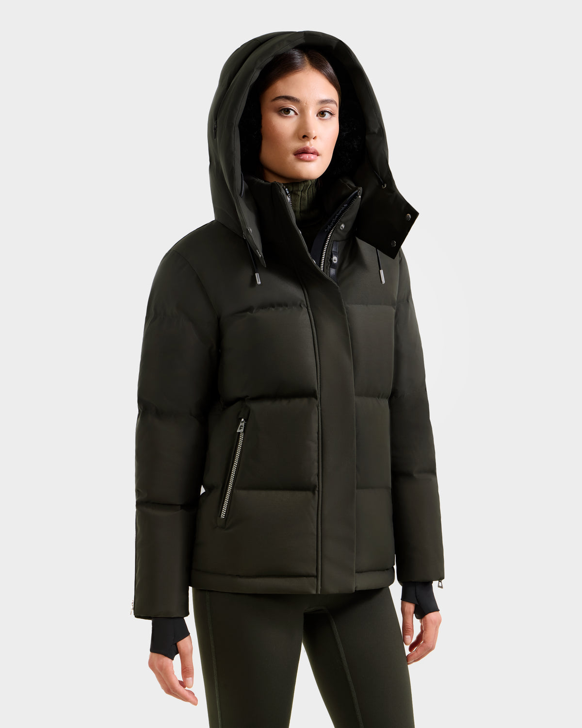 A woman wears the Rudsak JOON H - 8125586 Dark Olive women’s down jacket, featuring zippered pockets. She stands facing forward with her hands at her sides, paired with matching dark green pants for a stylish cold-weather look.