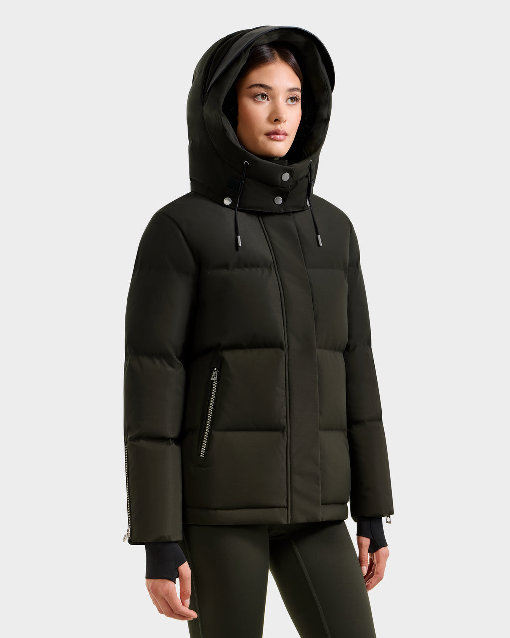 A woman wears the Rudsak JOON H - 8125586 women’s down jacket in dark olive, featuring a hood, high collar, and zippered pockets, standing against a plain white background.