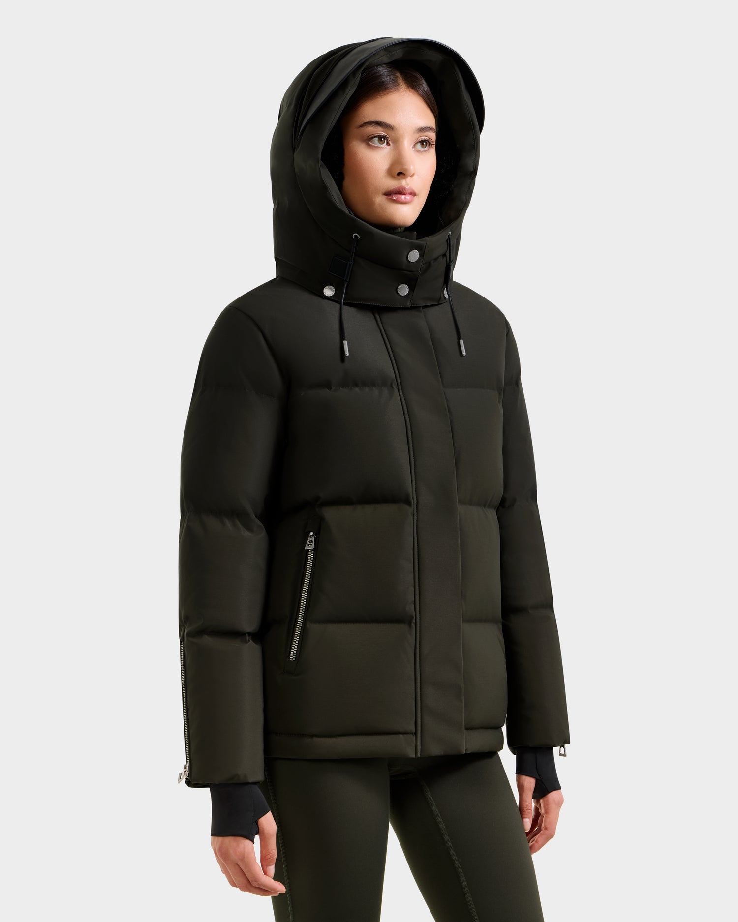 A woman wears the Rudsak JOON H - 8125586 women’s down jacket in dark olive, featuring a hood, high collar, and zippered pockets, standing against a plain white background.